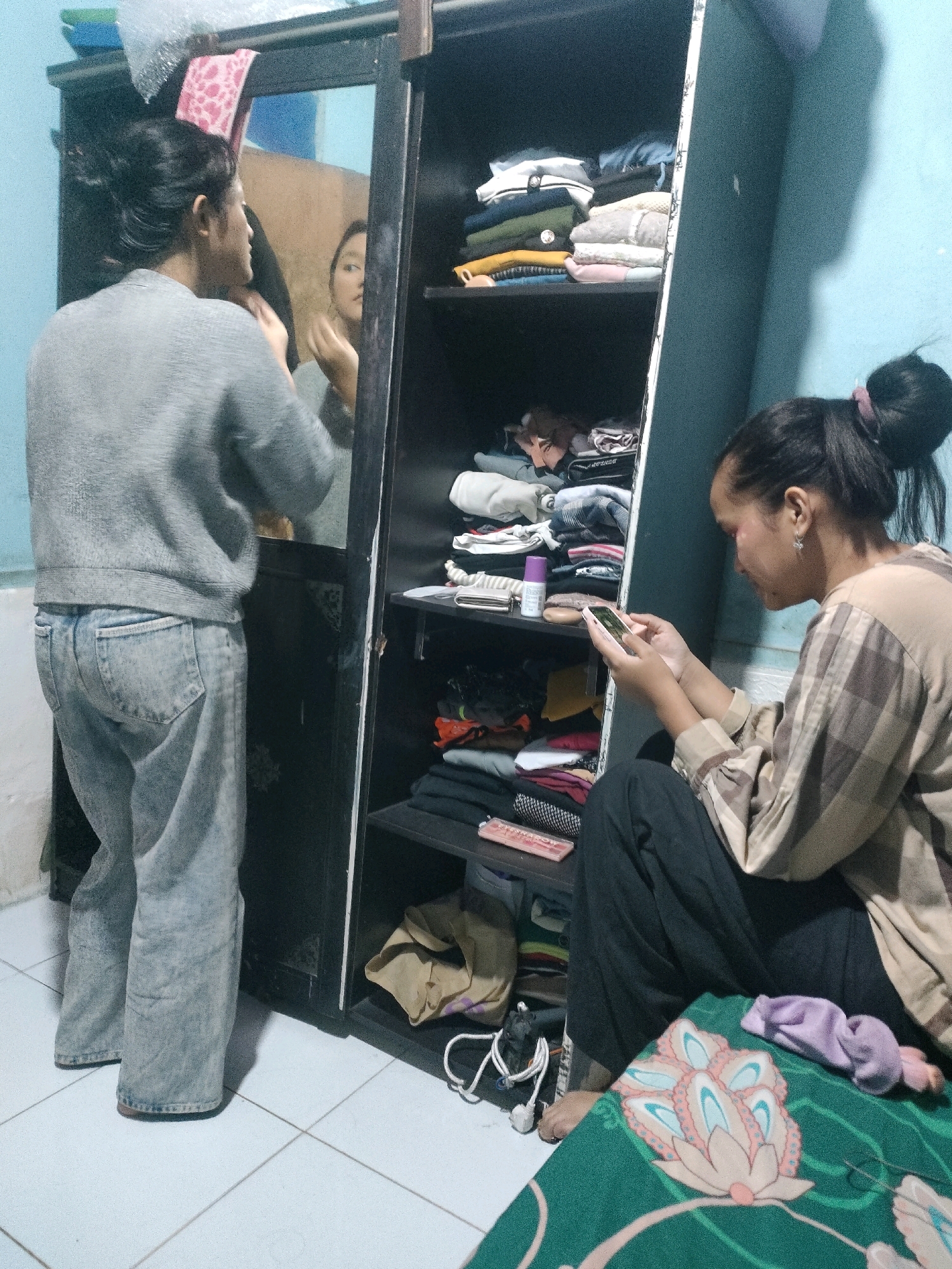 The image depicts an indoor scene, likely a bedroom or dressing area, in Pekanbaru, Indonesia. A person, seen from the back, is standing in front of a tall, dark wardrobe with mirrored doors. This individual is wearing a grey long-sleeved sweater and light-wash, loose-fitting jeans. They are looking into the mirror, with their reflection visible, and appear to be adjusting something near their face.
To the right of the wardrobe, another person is seated on the floor, leaning against the wardrobe. This individual is wearing a plaid long-sleeved shirt and dark pants. They are holding a smartphone in their hands and looking down at it, seemingly engaged with the device. The wardrobe is filled with neatly folded clothes on several shelves.
The environment appears to be a domestic setting, with a blue painted wall visible in the background and a tiled floor. A green blanket with a floral pattern is spread on the floor in the foreground, with a purple fabric item on top of it. Various household items, such as electrical cords and a beige fabric bag, are also present in the scene. The lighting suggests it is likely evening or dimly lit.