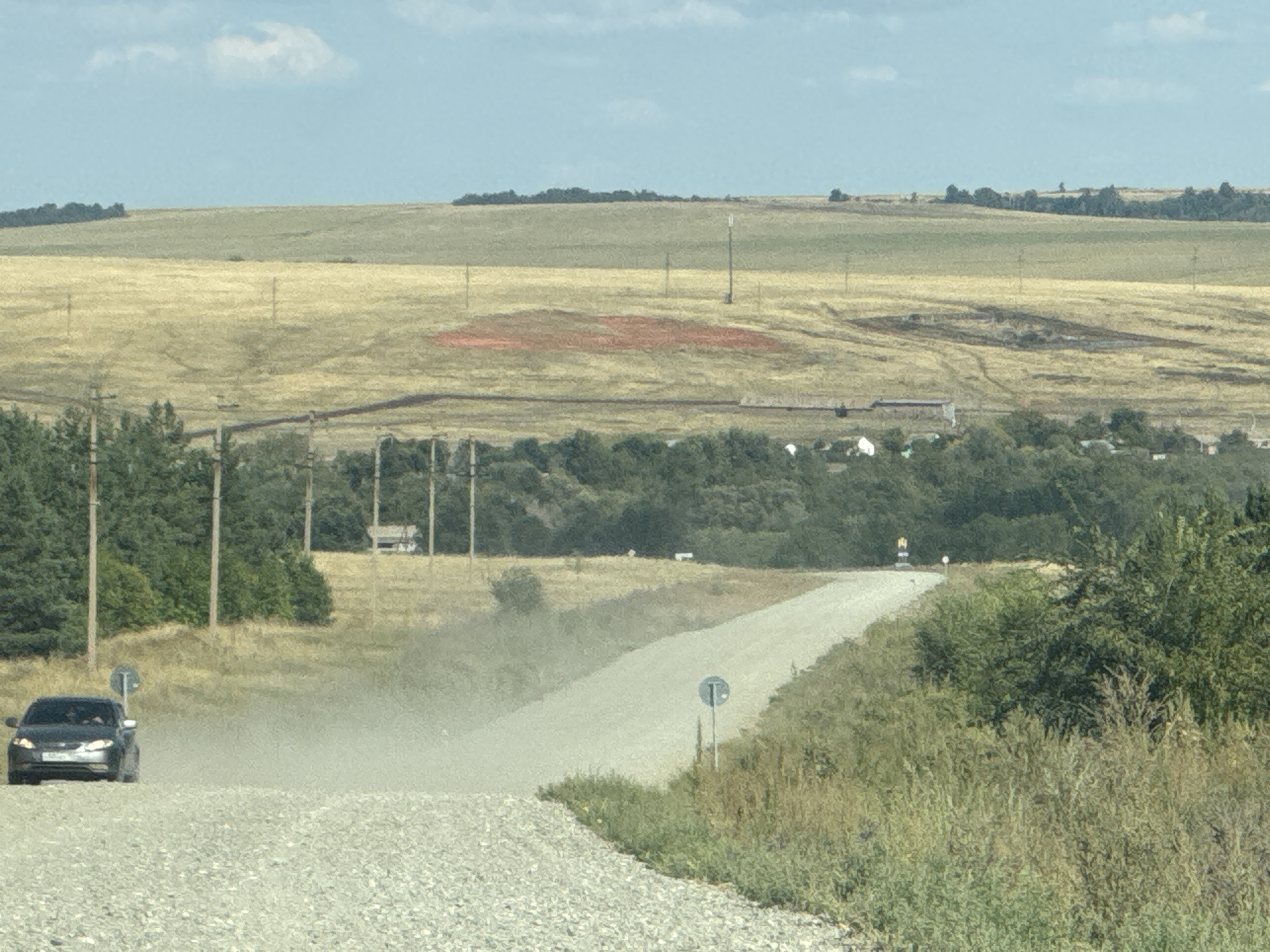 An eye-level, medium shot captures a rural landscape scene on a partly cloudy day. In the foreground, a gravel road curves to the right, kicking up dust as a dark sedan approaches from the left. The car, likely a passenger vehicle, is the main subject in motion. Beyond the road, a line of power poles stretches into the distance, interspersed with trees and fields. The fields are a mix of dry grass and some areas with a reddish hue, possibly indicating cultivated land or a natural phenomenon. In the far distance, a cluster of buildings suggests a small settlement. The overall impression is one of a remote, possibly agricultural, area in a rural setting, likely Васильевка, Russia, given the context.
