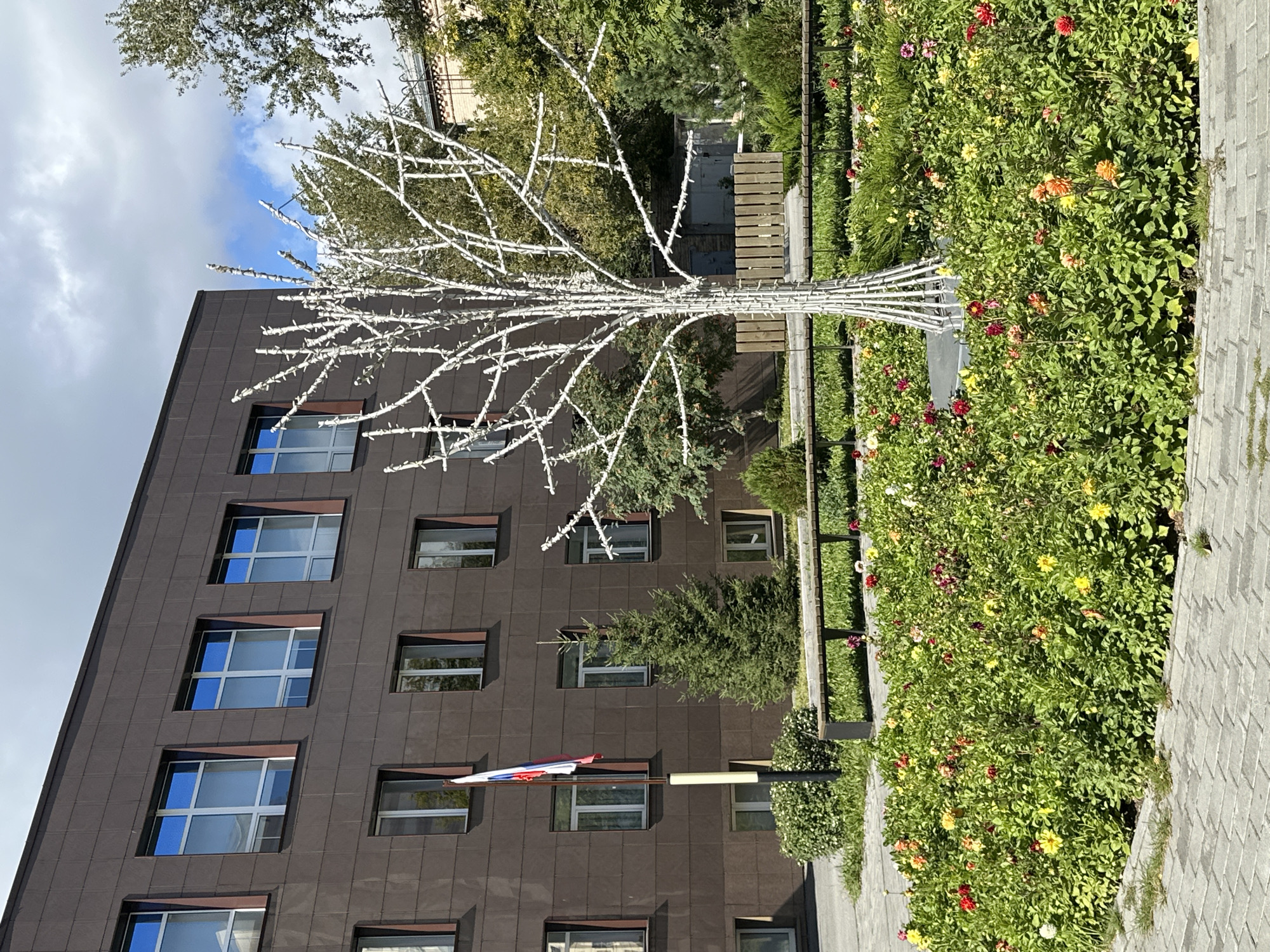 A white, artificial tree decorated with small lights stands in the center of a flower bed filled with red, yellow, and pink dahlias. The tree is positioned in front of a modern, dark brown building with several windows. To the left of the tree and building is a flagpole flying the Russian flag. A few evergreen trees are planted behind the flower bed, and a wooden bench is visible behind them. The ground in front of the flower bed is paved with gray bricks. The sky is partly cloudy.