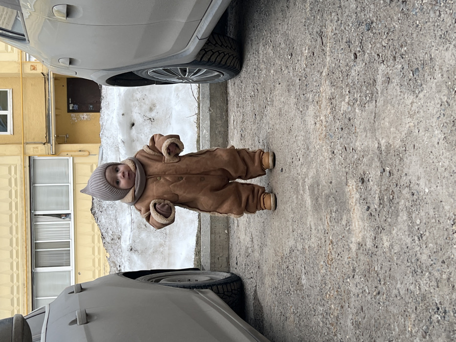 A forensic description of the scene depicts a young child, approximately a toddler, standing centrally in an outdoor, semi-urban environment in Noviy, Russia. The child is dressed in a warm, light brown, fur-lined full-body jumpsuit with visible buttons, a matching ribbed beanie with a pointed top, a coordinated neck warmer, and light brown shoes. The child faces forward, looking directly ahead, with arms slightly bent and hands clenched.
The setting appears to be a residential street or parking area. The ground surface consists of packed dirt and gravel. Two light-colored vehicles, possibly SUVs or sedans, flank the child, with only their lower front sections and tires visible on either side. Behind the child, a significant pile of melting, dirty snow and ice is present, indicating residual winter conditions. This snow pile rests beyond a concrete curb.
In the background, a multi-story residential building with a textured yellow facade is visible. Two windows are prominent on the lower floor; a faint, partial human figure is discernible looking out from the left window. A dark brown entrance door is located below the right window area. The overall lighting is overcast, producing soft, uniform illumination across the scene.