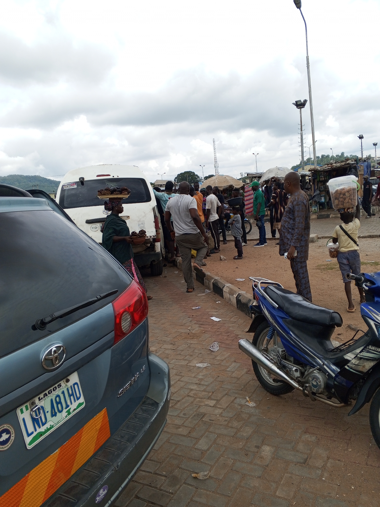 The image captures a bustling street scene in Lokoja, Nigeria, characterized by a mix of commerce, transportation, and pedestrian activity. A blue Toyota Sienna is parked on the left, its license plate reading LND-481HD, indicating registration in Lagos. Adjacent to it is a white van, partially visible, with a woman standing nearby, possibly selling food items displayed on a tray.
A crowd of people is gathered in the middle ground, including men in casual attire and some individuals in more formal, patterned clothing. Among them, a young boy carries a container of goods on his head, accompanied by a smaller pail. To the right, a blue motorcycle is parked.
The environment includes a brick-paved ground, a raised curb separating the roadway from the pedestrian area, and various streetlights illuminating the area. In the background, makeshift stalls and umbrellas suggest a marketplace setting. The sky is overcast, casting a diffused light over the scene.