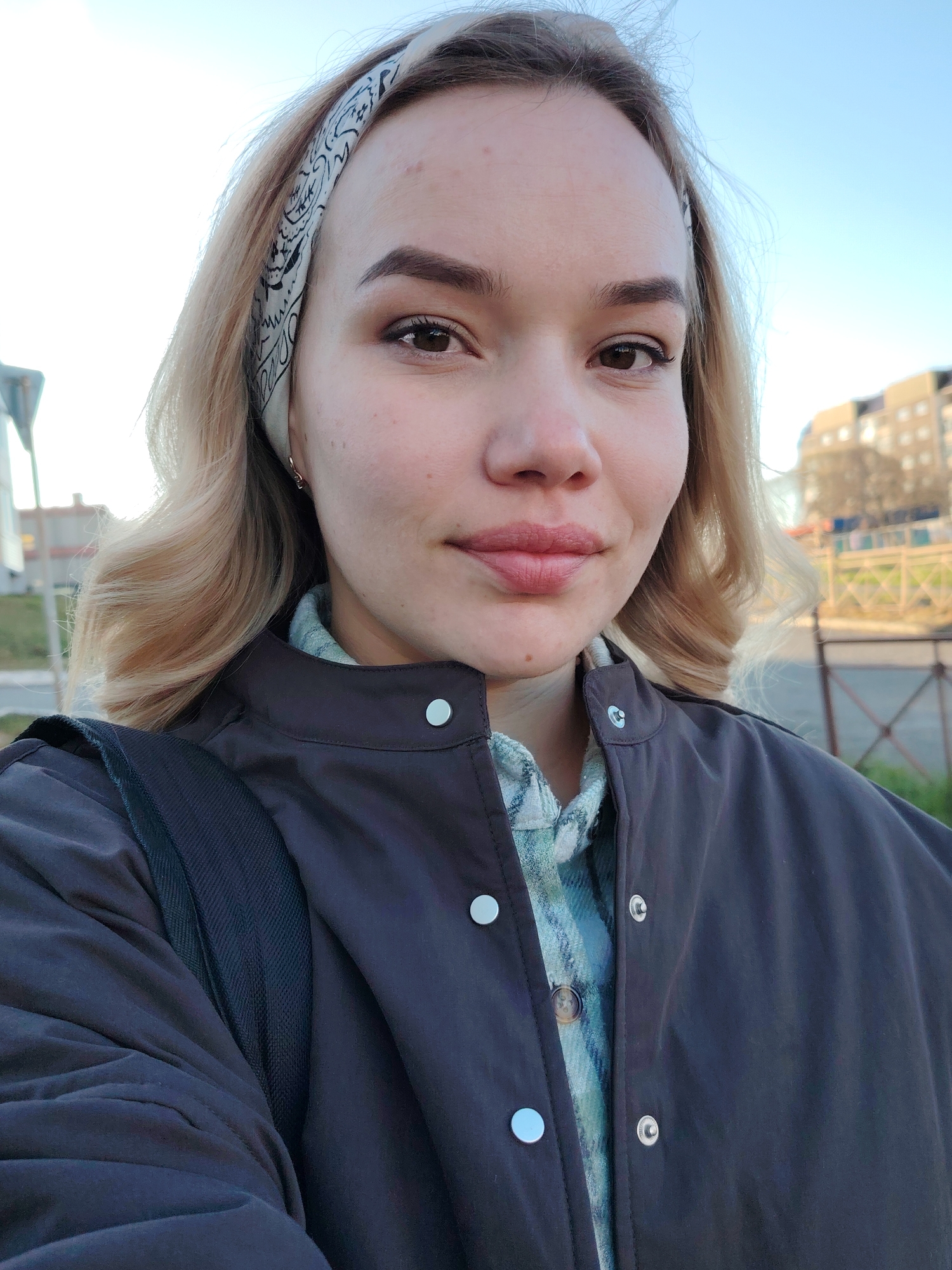 A young woman is taking a selfie outdoors. She has blonde, wavy hair and is wearing a patterned headscarf, a plaid shirt, and a dark jacket. A black backpack strap is visible on her left shoulder. The background includes a fence, a building, and some greenery. The image was taken in Markovskii, Russia.