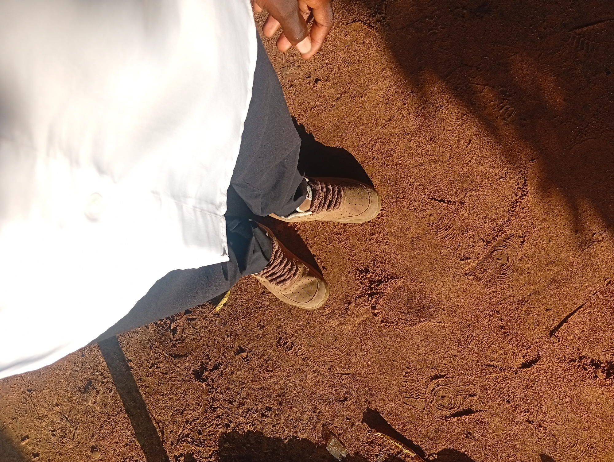 The image shows a person standing on reddish-brown dirt. The person is wearing dark pants and light brown sneakers with brown laces. Part of a white garment, possibly a lab coat or uniform, is visible on the left side of the frame, obscuring the rest of the person's body. The dirt ground has several shoe prints imprinted on it, indicating recent activity. Sunlight casts distinct shadows on the ground, suggesting it is daytime. The lighting and dry appearance of the dirt hint at a sunny, possibly warm, environment. The scene appears to be outdoors, in a location that is not paved. The visual cues of dirt, shadows, and footprints are consistent with a rural or less developed area. There is no visible text in the image. The context provided, Ukukwa, Nigeria, aligns with the visual characteristics of the scene, suggesting a possible outdoor gathering, work, or leisure activity in that location.