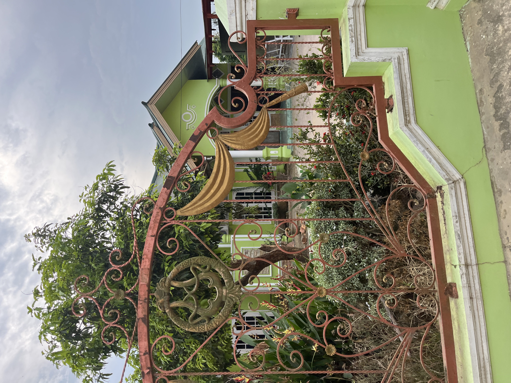 The image depicts the front of a bright green house, viewed from behind a red metal gate. The gate has intricate scrollwork and a circular emblem featuring a stylized, possibly mythical, creature. In front of the house, there is lush green foliage, including trees and flowering plants. The house itself has a green roof and white trim around the windows. A portion of a concrete walkway or patio is visible in the foreground. The sky is overcast. The location is identified as Tanahterban, Indonesia.