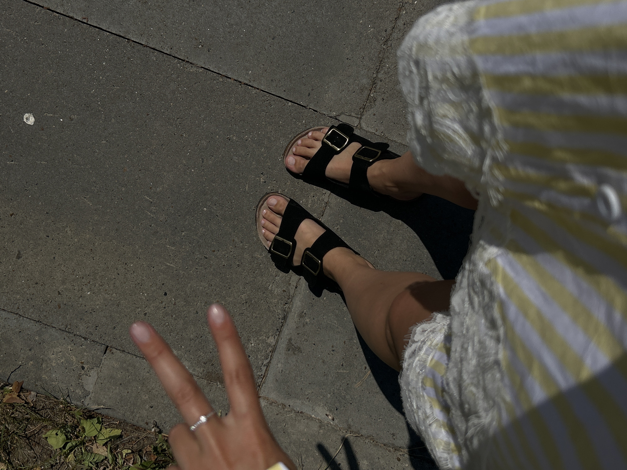 A high-angle, medium shot captures a person's legs and feet as they stand on a cracked concrete sidewalk. The person is wearing black Birkenstock-style sandals with gold buckles and a white and yellow striped garment. A hand with a ring on the index finger is visible in the upper left corner, forming a peace sign. The ground is a mix of concrete and dry grass, and a shadow is cast on the sidewalk by the person's body and the surrounding environment. The scene suggests an outdoor setting, possibly in a park or urban area.