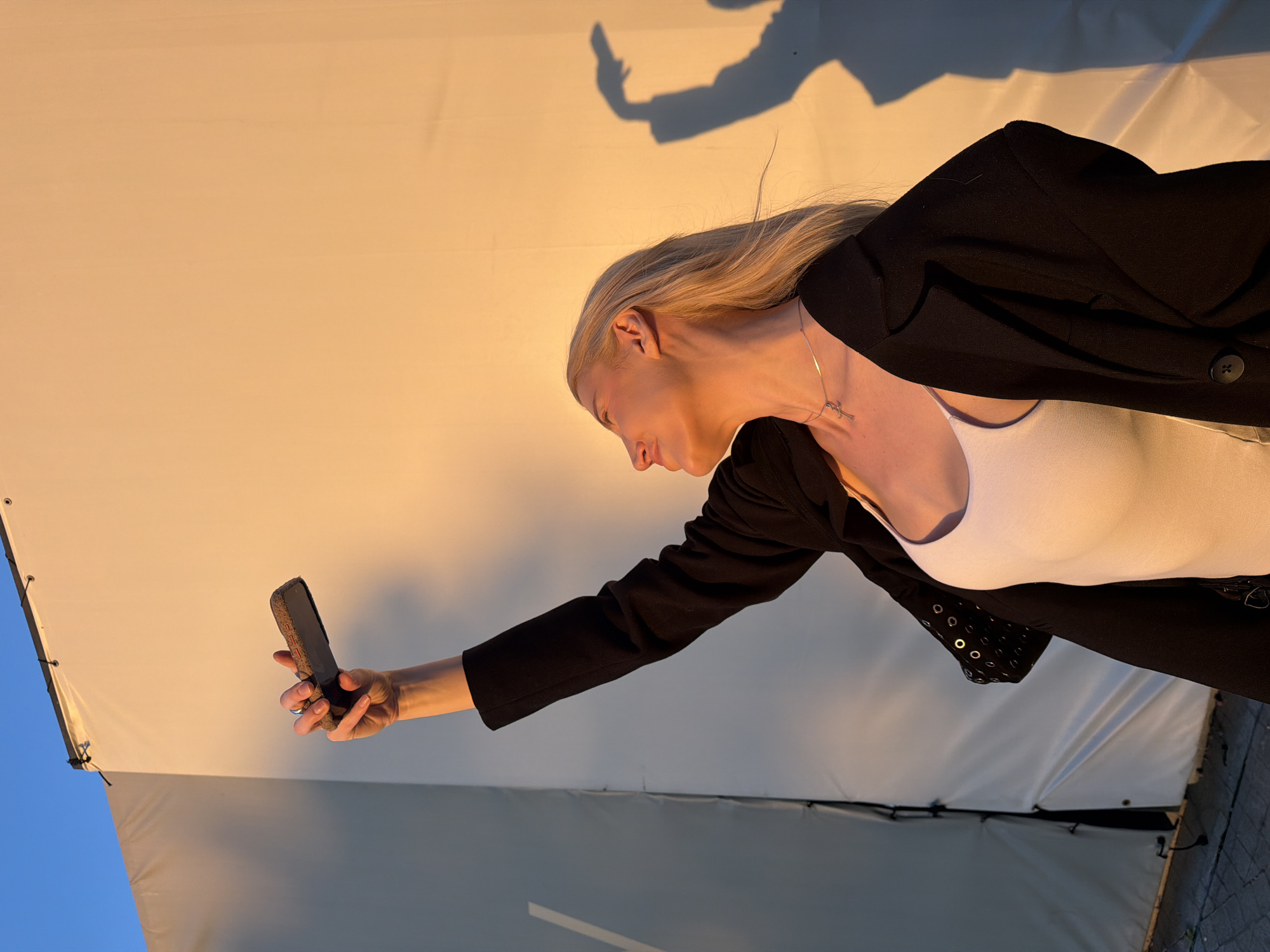 A woman with fair skin and long blonde hair is holding a mobile phone. She is wearing a white top and a black jacket. The photo appears to be taken outdoors near a white backdrop with a shadow projected onto it. The location is the city of Belgrade, Serbia.