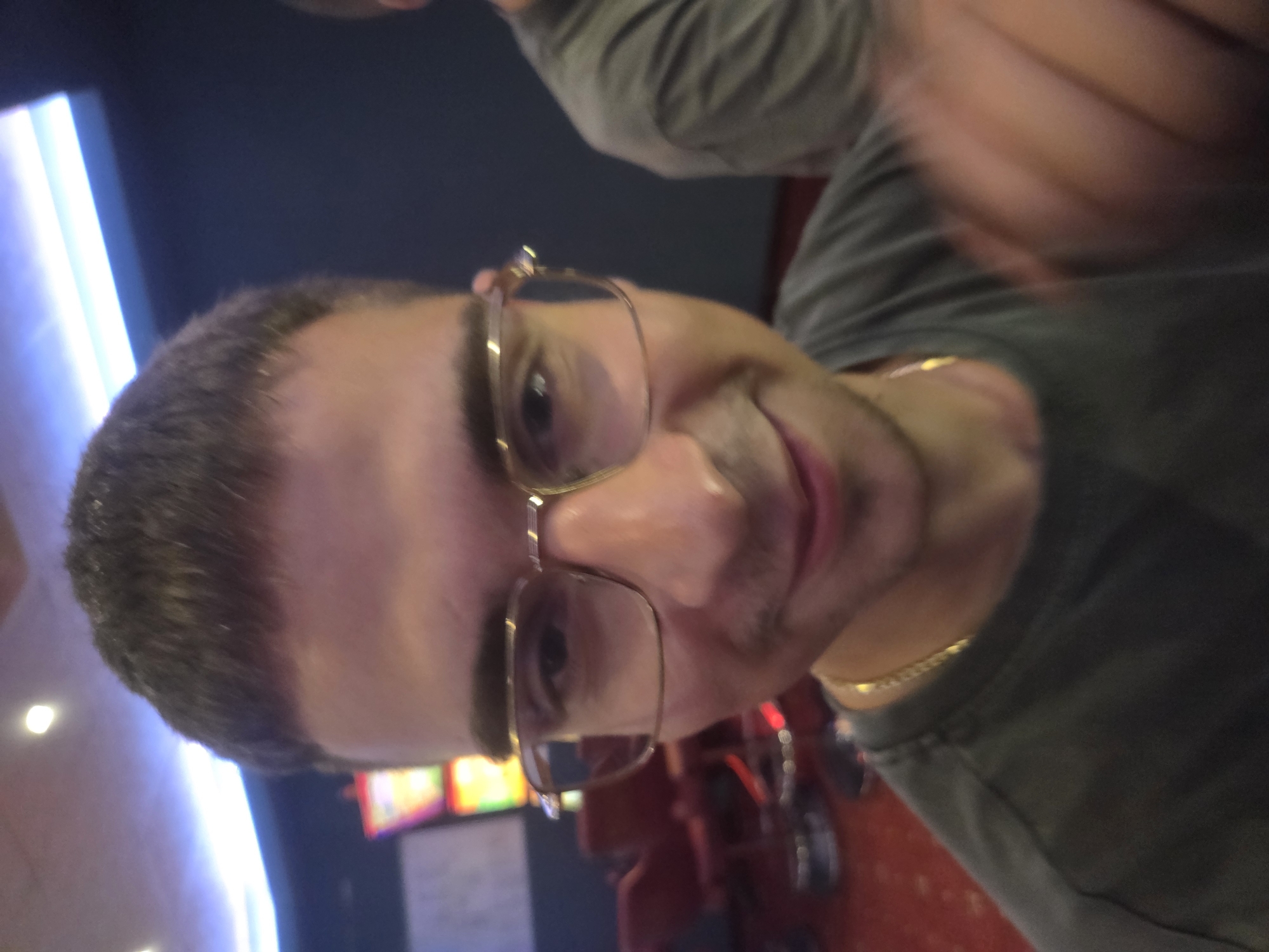 The image shows a close-up, selfie-style photograph of a young man wearing glasses and a dark t-shirt. He has short, dark hair and is looking at the camera with a slight smile. A gold chain is visible around his neck. In the background, red chairs and what appear to be slot machines are visible, suggesting the location could be a casino or entertainment venue. The lighting in the background is dim with some bright, overhead lights. The image does not provide any information about the city of Beloslav, Bulgaria.