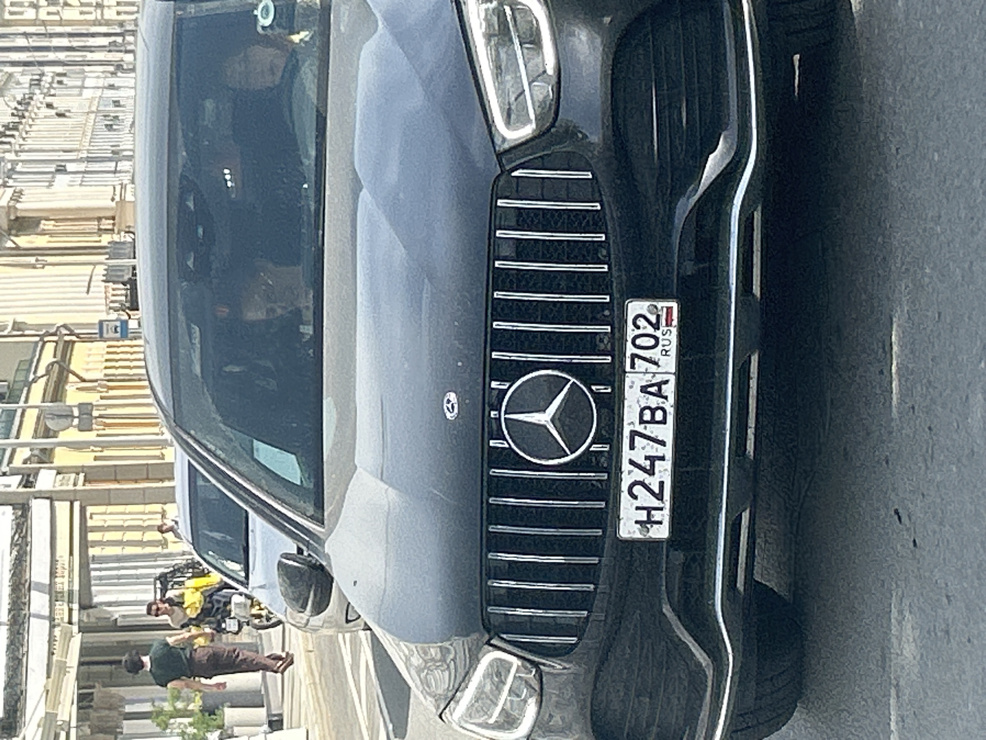 The image shows the front of a black Mercedes-Benz car with a Russian license plate H247BA 702. The car is parked on a paved surface, possibly a street. In the background, there are people walking and a street cleaning vehicle. The location appears to be an urban area.