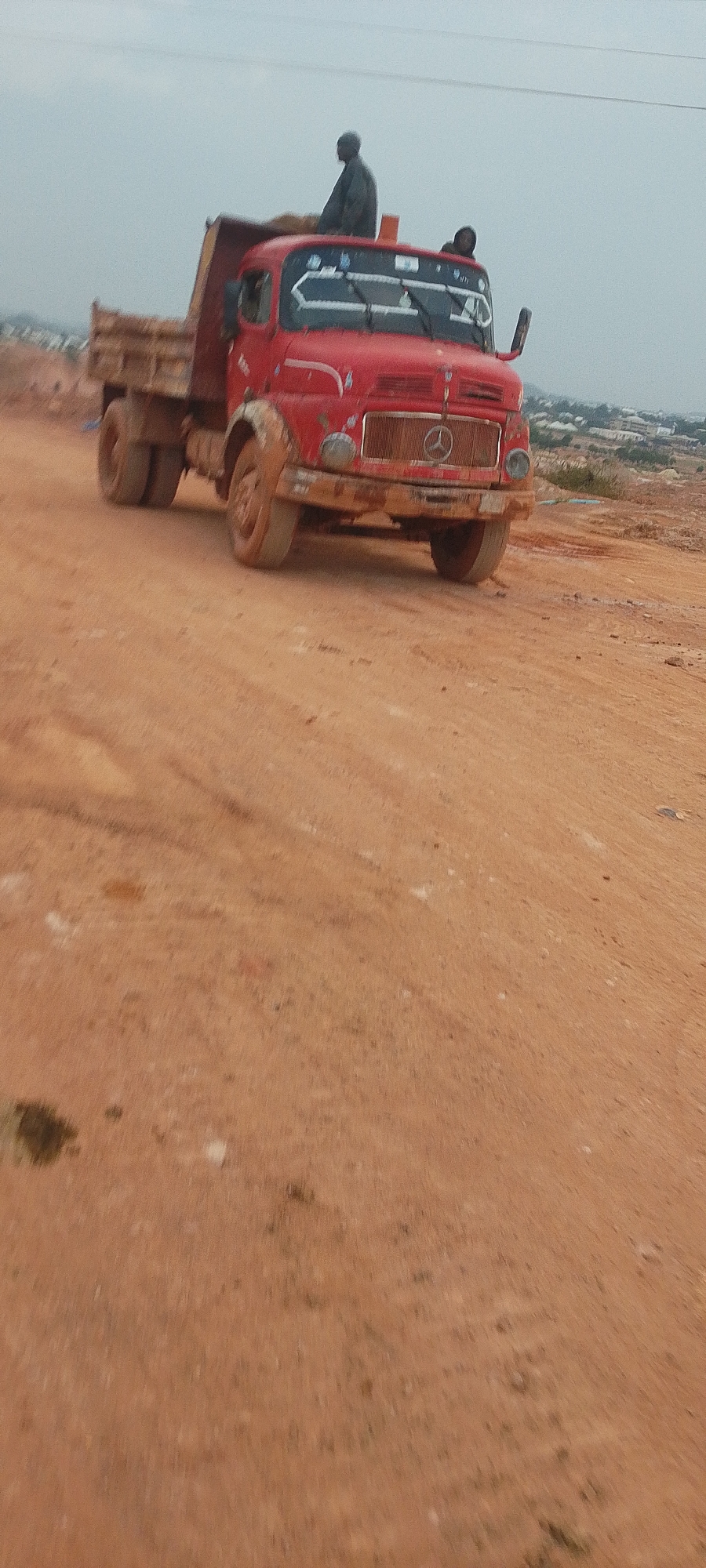 The image shows a red Mercedes-Benz dump truck with two people in the cab and one standing in the back, likely a construction site in Reifild-Ist, Nigeria. The truck is covered in mud, suggesting it is actively engaged in construction or hauling materials. The ground is dirt and sand, consistent with an undeveloped or under-construction area. The sky is overcast, and the background shows a hazy view of a town or city. The truck appears to be moving or has recently stopped on a dirt road.