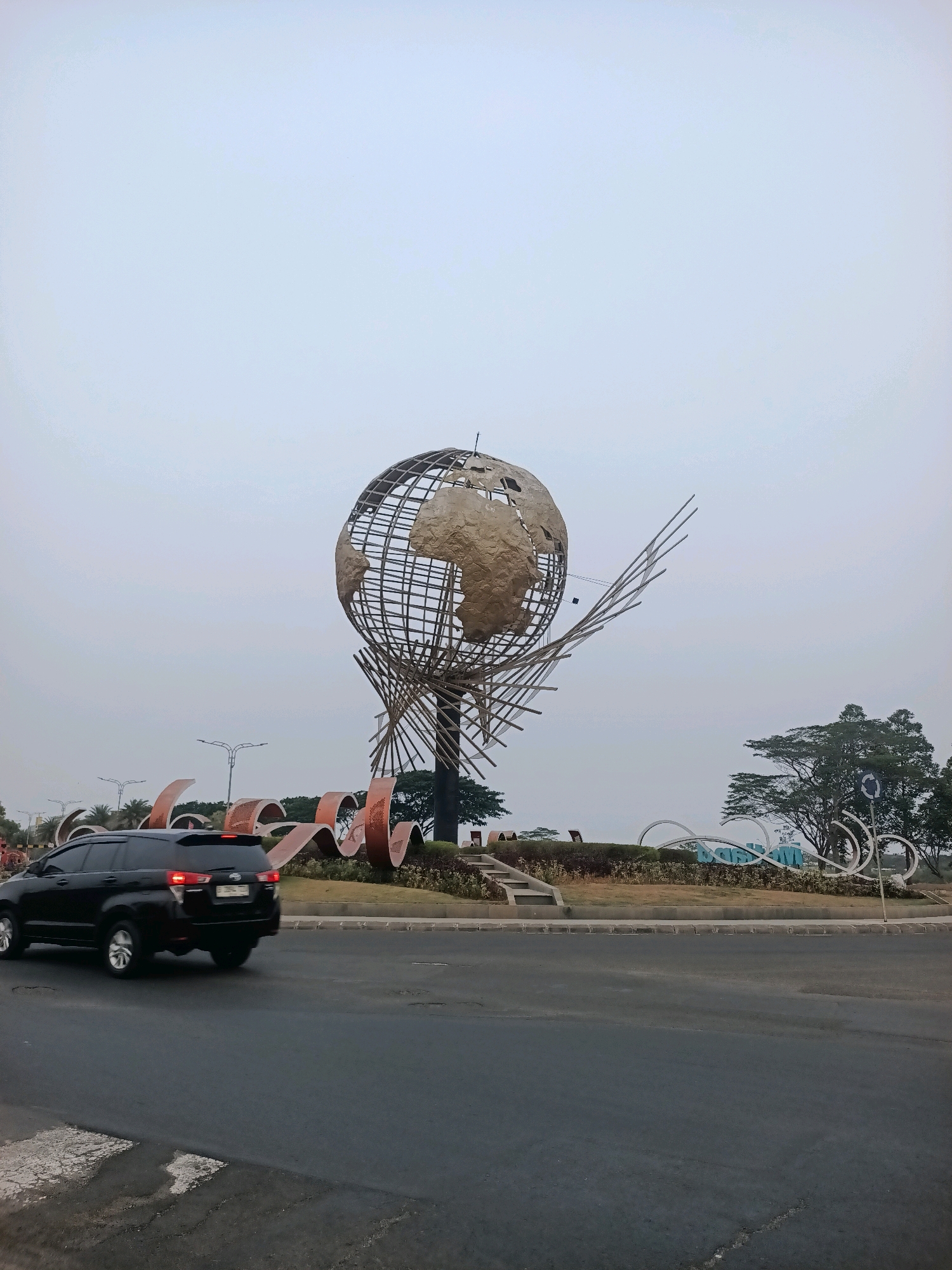 A black SUV drives on a road in front of a large, artistic globe sculpture. The sculpture is made of metal, with a wireframe sphere that has sections of what appears to be a gold-colored material representing landmasses. The globe is supported by a framework of wooden-like beams arranged in a dynamic, spiraling pattern. In front of the globe, large, curved, copper-colored structures form part of the artistic installation. In the background, there is a large sign with white, abstract lettering, partially obscured by trees and shrubs. The sky is overcast, creating a soft, diffused light. The road is asphalt, with white lane markings and some visible cracks. The overall scene suggests an urban environment, likely a public space or monument in Telagamurni, Indonesia, given the presence of the specific sculpture.