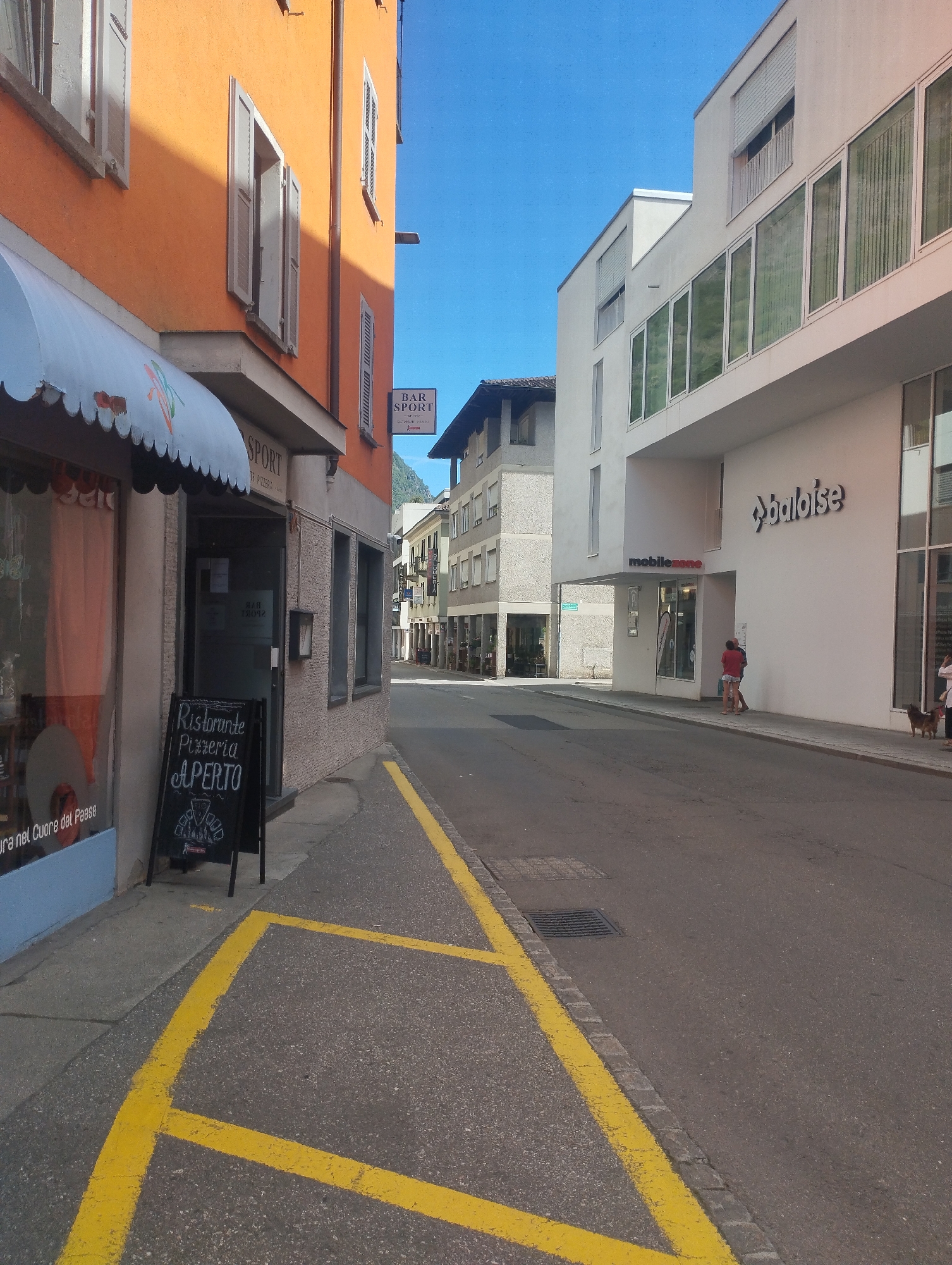 The image depicts a street scene in a European-style town under a clear blue sky. The viewpoint is from street level, looking down a road that curves to the right.
On the left side of the street, an orange building dominates the foreground. This building features multiple windows, some with closed shutters and others with open shutters. A white awning extends over a storefront on this building. A black sandwich board sign stands on the sidewalk in front of the storefront, displaying "Ristorante Pizzeria APERTO" with a drawing of a man in a hat. A sign for "SPORT" is visible on the building.
Across the street, a modern white building with large glass windows runs along the right side of the road. This building has a sign that reads "baloise" and another smaller sign that says "mobilezone".
Further down the street, other buildings with balconies and awnings are visible. The road itself is asphalt, marked with yellow lines. A drain grate is visible in the road near the curb.
Several people are present in the scene:
Two individuals, one in a red top and the other in a blue top, stand on the sidewalk in front of the "mobilezone" store.
A woman is walking a dog on the sidewalk in front of the "baloise" building.
The overall impression is a sunny day in a town with a mix of older and modern architecture.