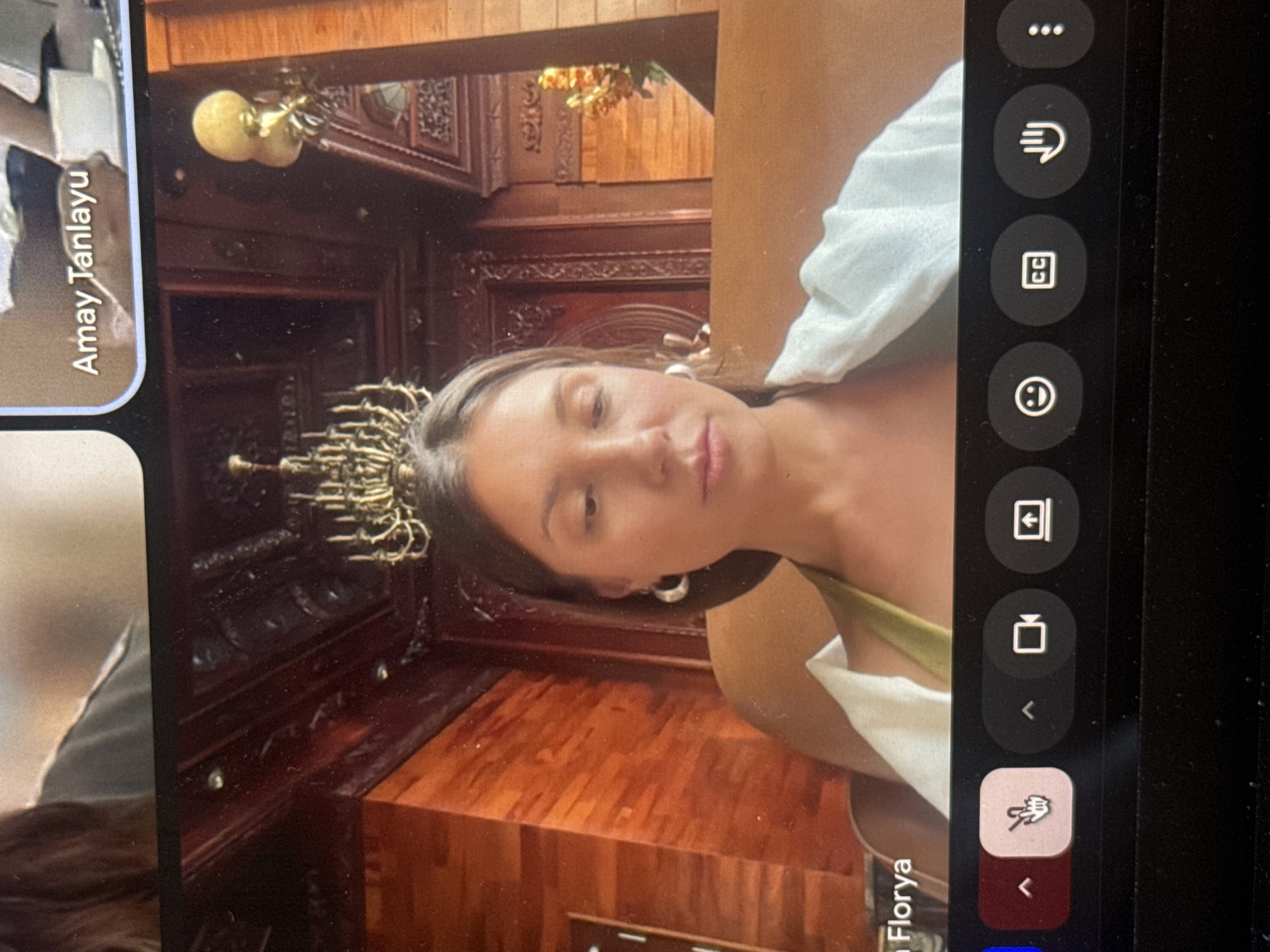A photo depicts a video call on a tablet device. Two participants are visible: one labeled "Amay Tanlayu" and another "Florya". The background behind "Florya" features ornate wooden furniture and what appears to be a chandelier. Given the architectural style and the person labeled as Florya, it is possible that the location is in Đà Nẵng, Vietnam, within a building or residence featuring traditional Vietnamese interior design, although this cannot be confirmed. The quality of the photograph is poor, and the angle of the tablet is not straight. The tablet's video call interface is visible, including interaction buttons on the side.