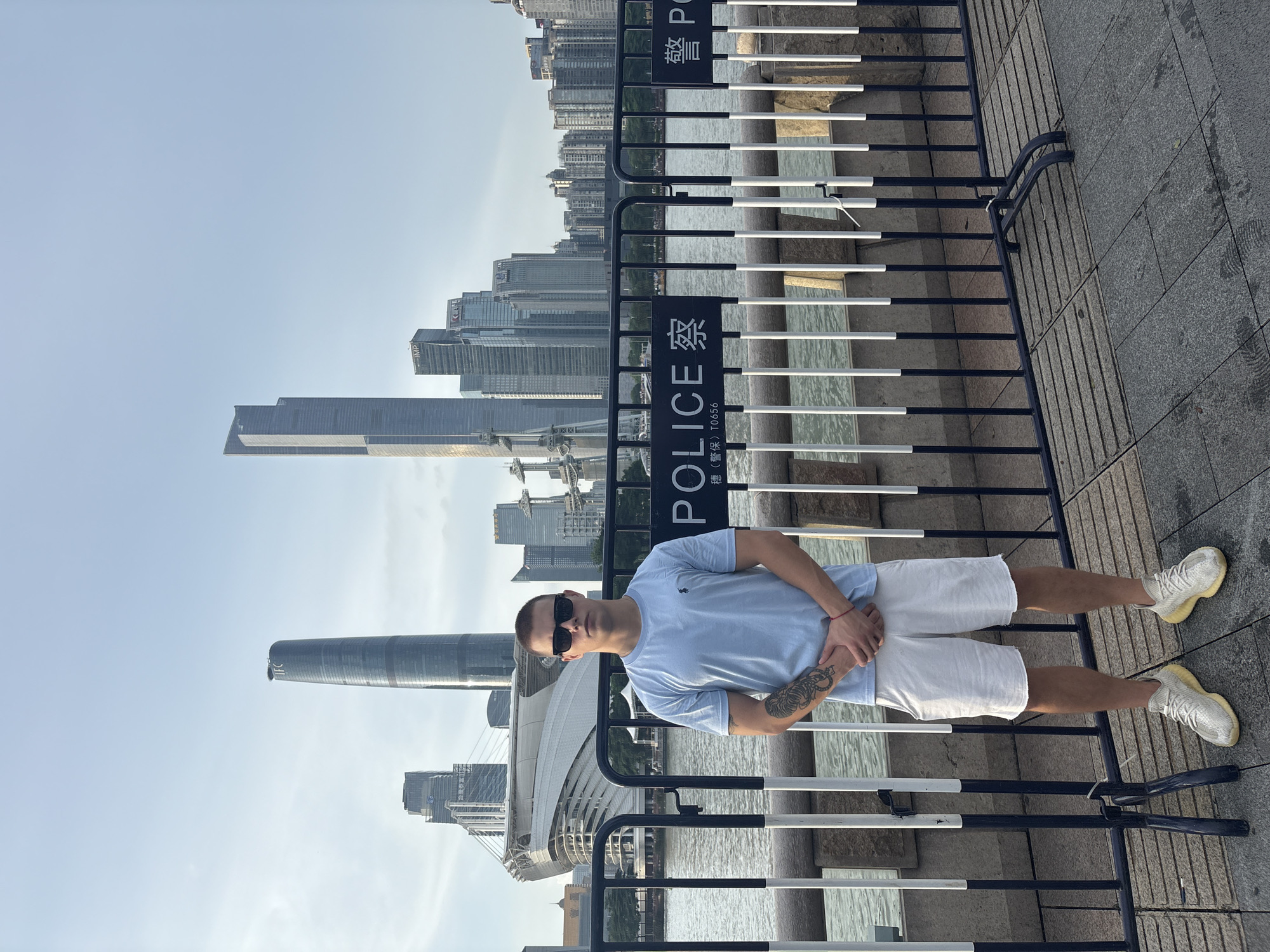 A young man with short hair and sunglasses stands in front of a metal barrier. He is wearing a light blue t-shirt, white shorts, and light-colored sneakers. He has a tattoo on his left arm. The barrier has "POLICE" written on it in English and Chinese.
Behind the barrier is a body of water, and across the water, a cityscape with numerous tall buildings is visible. The sky is clear and bright, suggesting it is daytime. The scene appears to be outdoors, possibly along a waterfront promenade or viewing area. The orientation of the image is vertical.