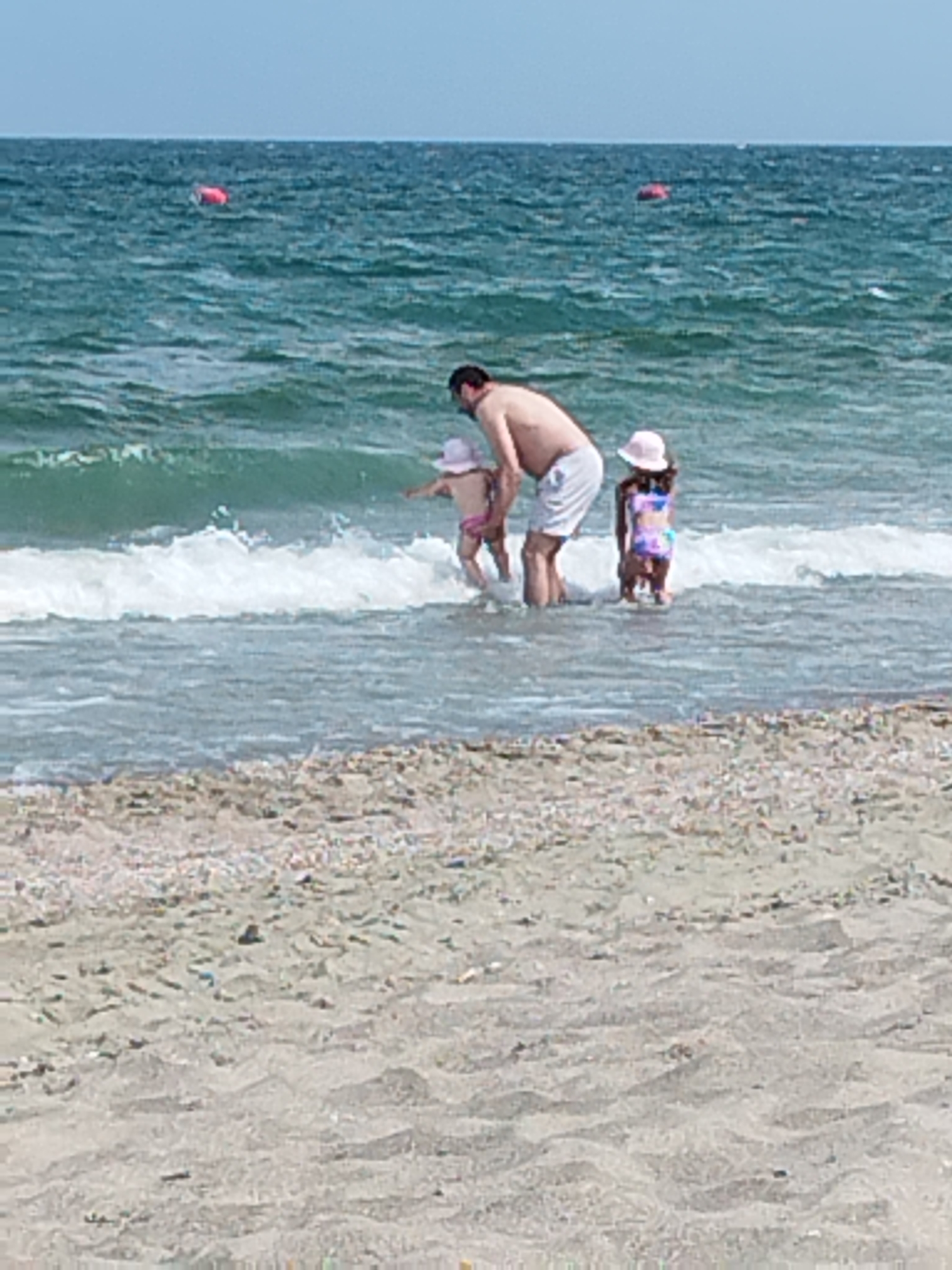 An adult male and two young children are standing in the shallow water at a beach. The male is wearing white shorts and the children are wearing swimsuits and hats. The sea is a greenish-blue and there are gentle waves. In the background, two red buoys are visible in the water. The foreground is a sandy beach with small shells. This scene is consistent with a location along the coast of Constanța, Romania.