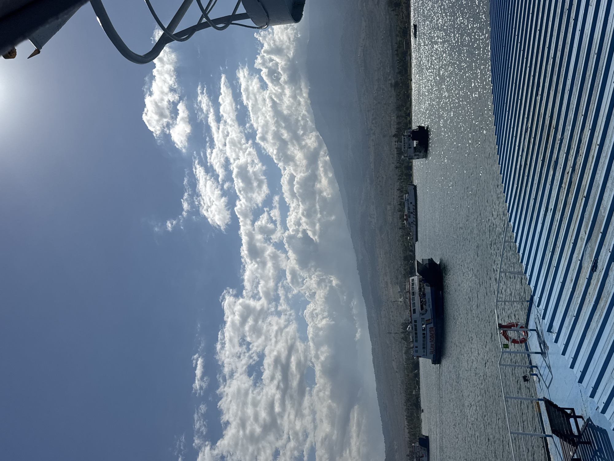 The image depicts a scene from the waters of Serunimumbul, Indonesia. The foreground shows the deck of a ferry with blue corrugated metal roofing and a white railing. A wooden bench and a red life preserver are visible on the deck. In the middle ground, several ferries are navigating the water, with one prominent ferry in the center displaying the text "Big Love Indonesia" on its side. The background features a hazy, mountainous landscape with a line of trees along the coast. The sky is a bright blue with scattered white clouds, and the sun is visible, creating a glare on the water. The overall impression is a bright, sunny day at sea with ferry traffic.