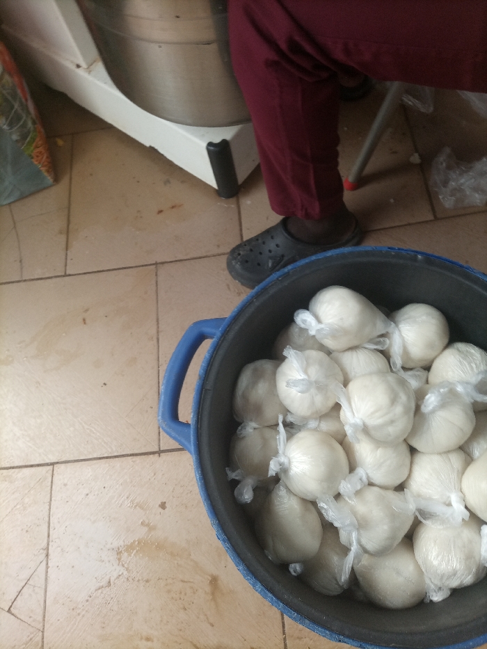 In Opete, Nigeria, an indoor scene displays a dark blue plastic bucket in the foreground, filled with approximately two dozen spherical, off-white food portions, each individually wrapped in clear plastic and secured with a knot. To the upper right, the lower portion of a person's legs is visible, clad in maroon trousers and dark open-backed clogs. This individual is positioned near a large, white industrial-style machine with a metallic bowl, visible in the upper left background. The machine rests on a white base with black protective feet. A metal stool leg with a red cap is partially visible behind the person's legs. The floor is tiled with light-colored, possibly beige, square tiles, exhibiting minor stains and grout lines. A patterned bag is partially visible on the far left.