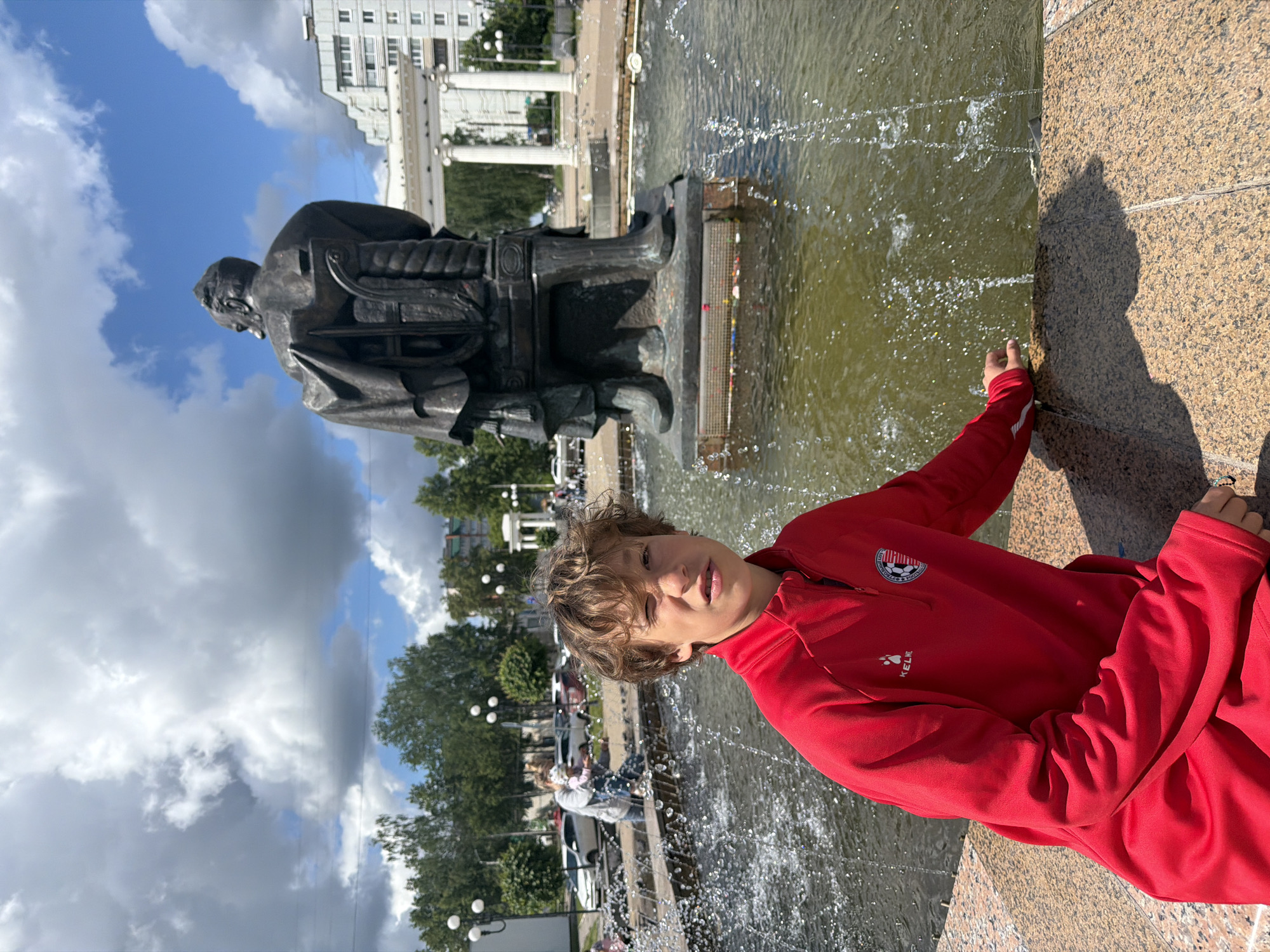 A young person is positioned beside a water fountain with a statue in Chaikovsky, Russia. The statue, a dark metal sculpture of a seated man, stands on a raised platform in the center of the fountain. The fountain water is slightly murky, with water jets arcing into the pool. The young person is leaning against the side of the fountain, wearing a red sports jacket and pants. A building with a white facade and multiple windows is visible in the background, along with trees and parked cars. The sky is partly cloudy with both dark and bright clouds.