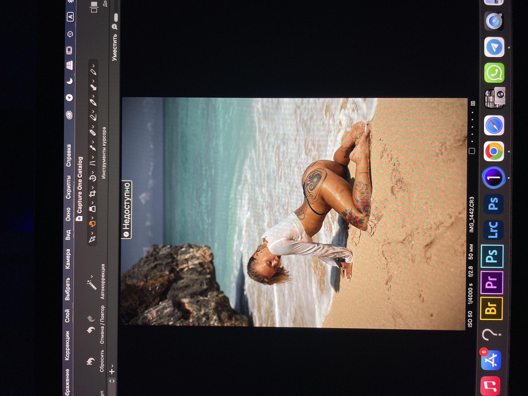 A rotated image of a person with tattoos on a beach, possibly in a tropical location, is displayed on a screen. The person is kneeling in the sand near the water. A rocky outcrop is visible in the background to the left. Various editing software tools are visible on the screen, indicating that the image is being edited. The file name "IMG_1442.CR3" suggests it's a photo file type. The screen is likely an iPad based on the software displayed. There's no evidence to suggest this photo was taken in Kazan, Russia.