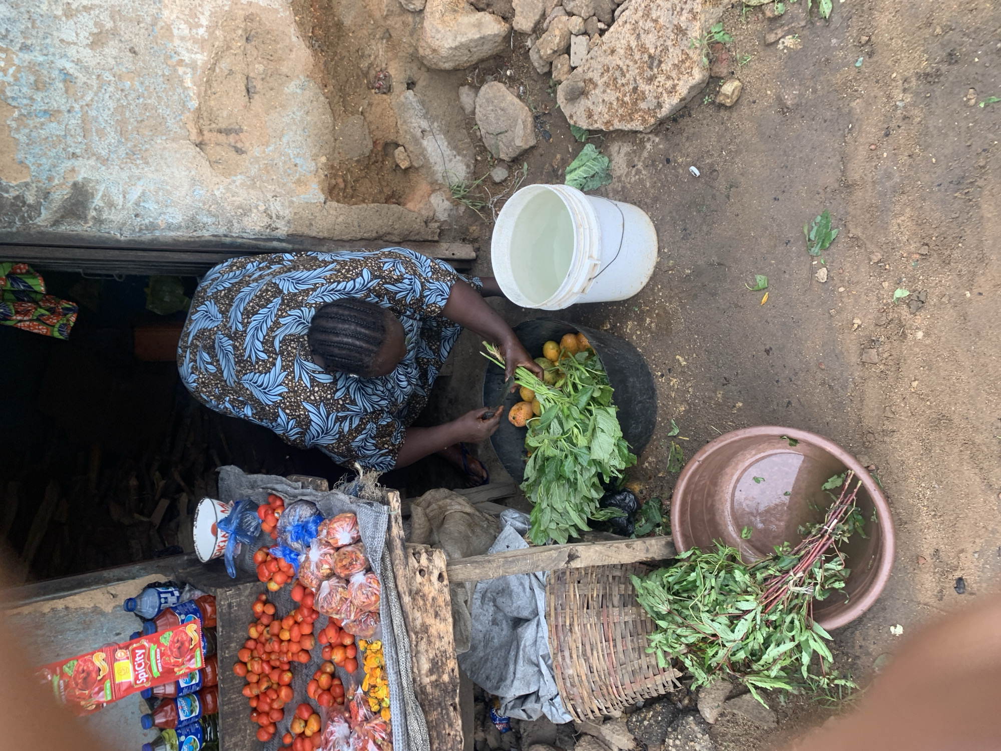 An overhead shot depicts a woman with braided hair, wearing a blue and brown patterned top, standing near a vendor stall in what appears to be Jos, Nigeria. The stall displays tomatoes, bottled drinks, and packets of what may be tomato paste. The woman is seen handling leafy green vegetables and yellowish-orange fruits, likely tomatoes or peppers, near a white bucket filled with water. Next to her are a large black plastic tub, a basket, and a brown plastic basin containing water and more leafy vegetables. The ground around the stall appears to be dirt and rock. A concrete structure forms part of the backdrop.