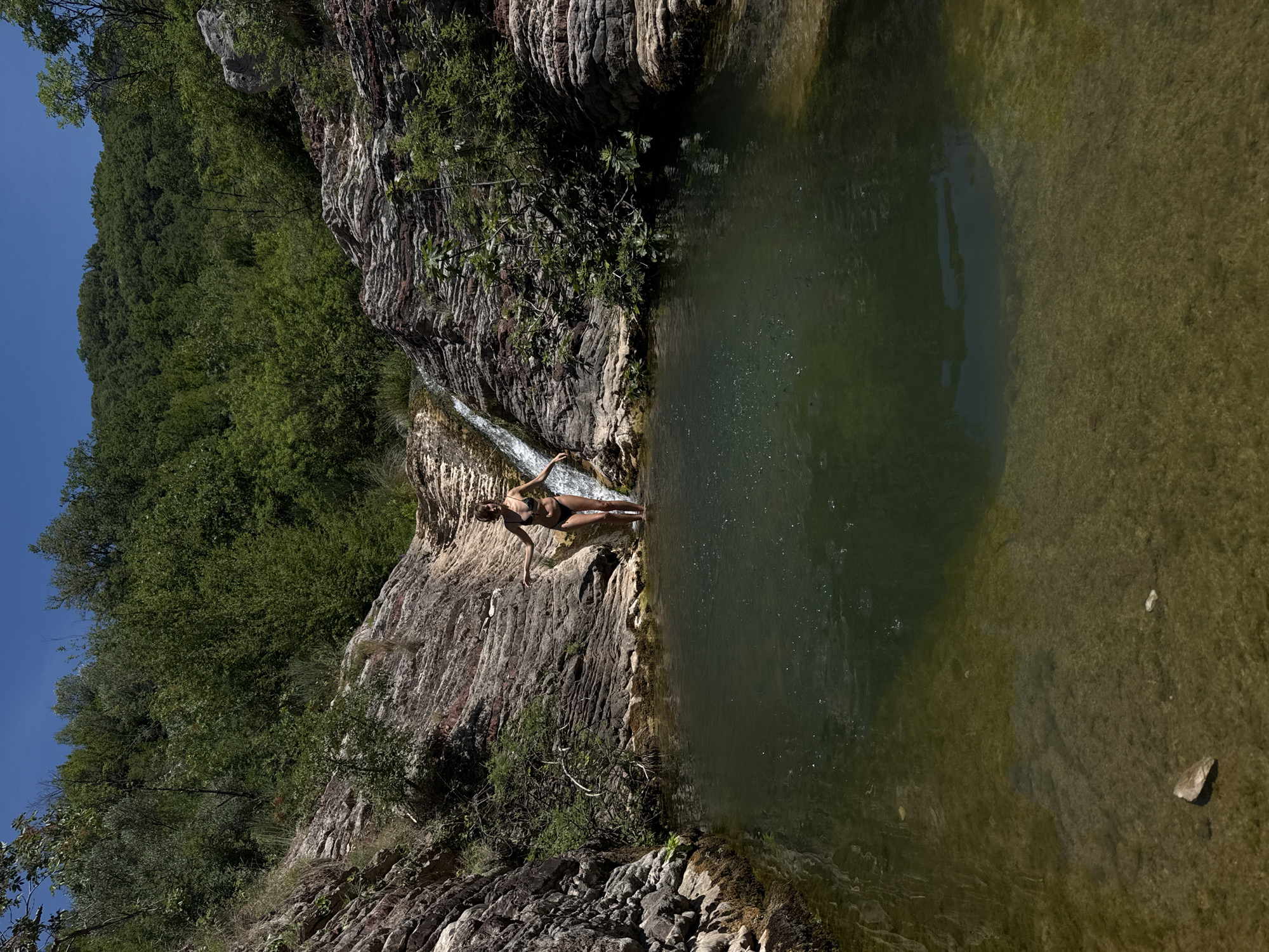 A full shot shows a person in a bikini standing in a shallow pool of water at the base of a small waterfall. The woman is facing the camera with her arms outstretched. The water is dark green and clear enough to see the rocky bottom. The waterfall cascades down a rocky cliff face. The surrounding environment is lush and green, with trees and vegetation covering the slopes. The sky is a clear blue. The scene appears to be a natural outdoor setting.