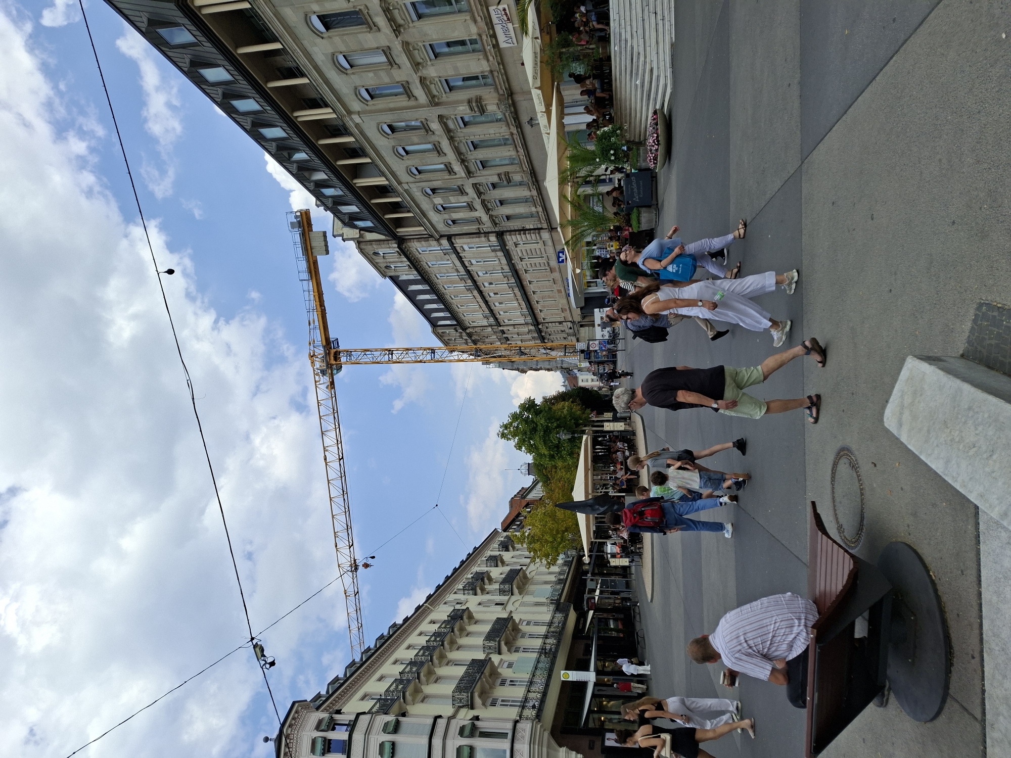 The image captures a street scene in Baden-Baden, Germany, likely in the city center. A large yellow construction crane dominates the upper portion of the frame, spanning across the sky. The street is paved with gray and darker gray square tiles, indicating a pedestrianized area or a public square.
On the left side of the street, a multi-story building with ornate balconies and a light-colored facade is visible. Further down, a building with shops at street level is present, featuring awnings and signage. A red "ALDI" sign is partially visible.
On the right side of the street, another multi-story building with a darker, mansard-style roof and numerous windows stands prominently. This building appears to house commercial establishments on the ground floor, with outdoor seating areas shaded by umbrellas. A sign reads "Amadeus Restaurant".
Several people are present in the scene. In the foreground, a man in a striped shirt sits on a distinctive, curved wooden bench. A family group, including adults and children, is crossing the street. Other pedestrians are walking in various directions.
A black sculpture stands in the middle of the street, near an outdoor seating area. Overhead power lines and cables crisscross the sky. The sky is partly cloudy with patches of blue. The overall impression is of a bustling urban environment with ongoing construction.