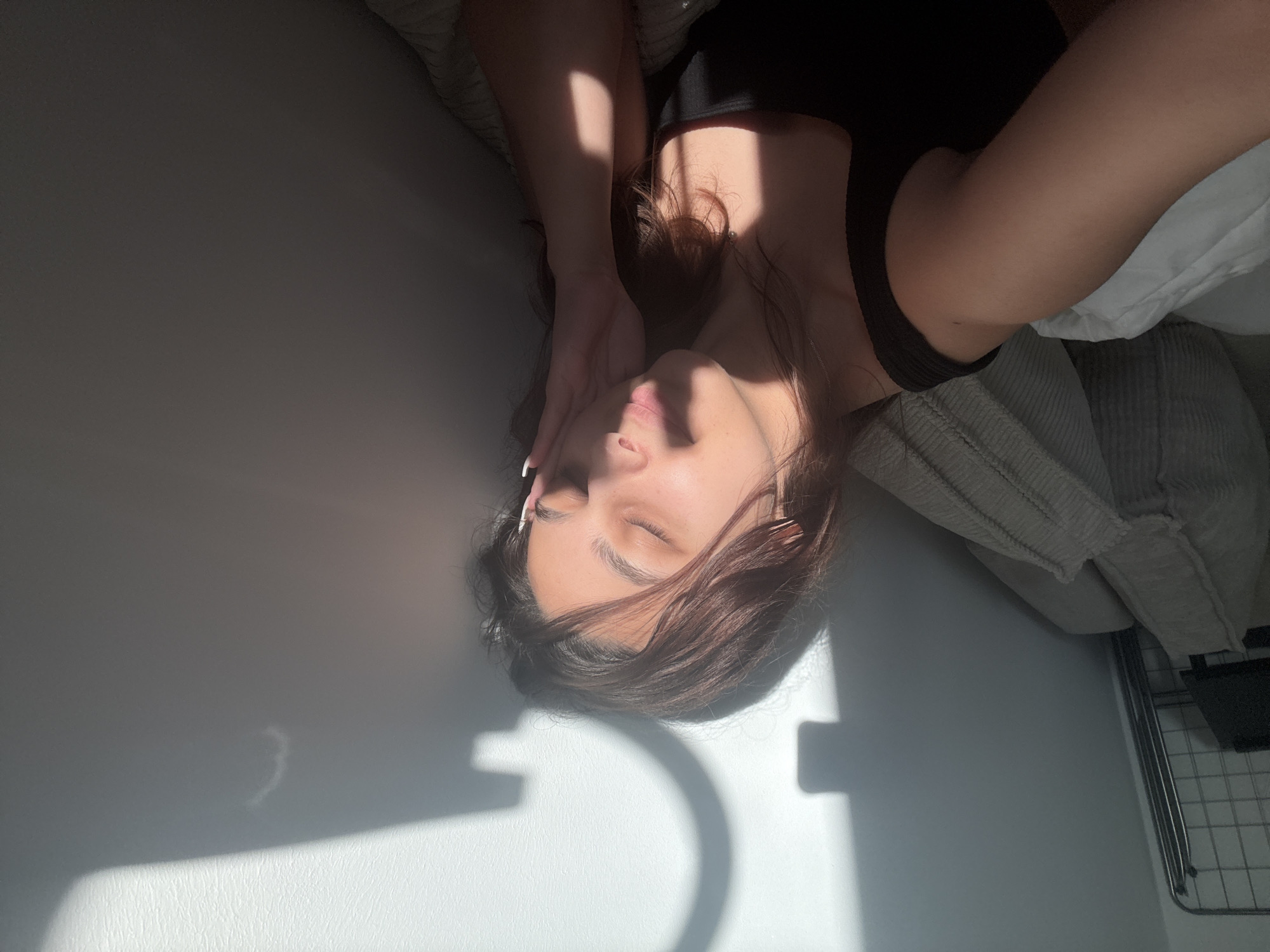 A young woman lies on a gray couch or bed with white pillows, wearing a black top. Her head is resting on her hand, with her eyes closed. Harsh sunlight casts shadows on her face and the wall behind her. A metal object, possibly a shelf, is visible in the lower right corner. The location, Mönchengladbach, Germany, is not evident from the image.
