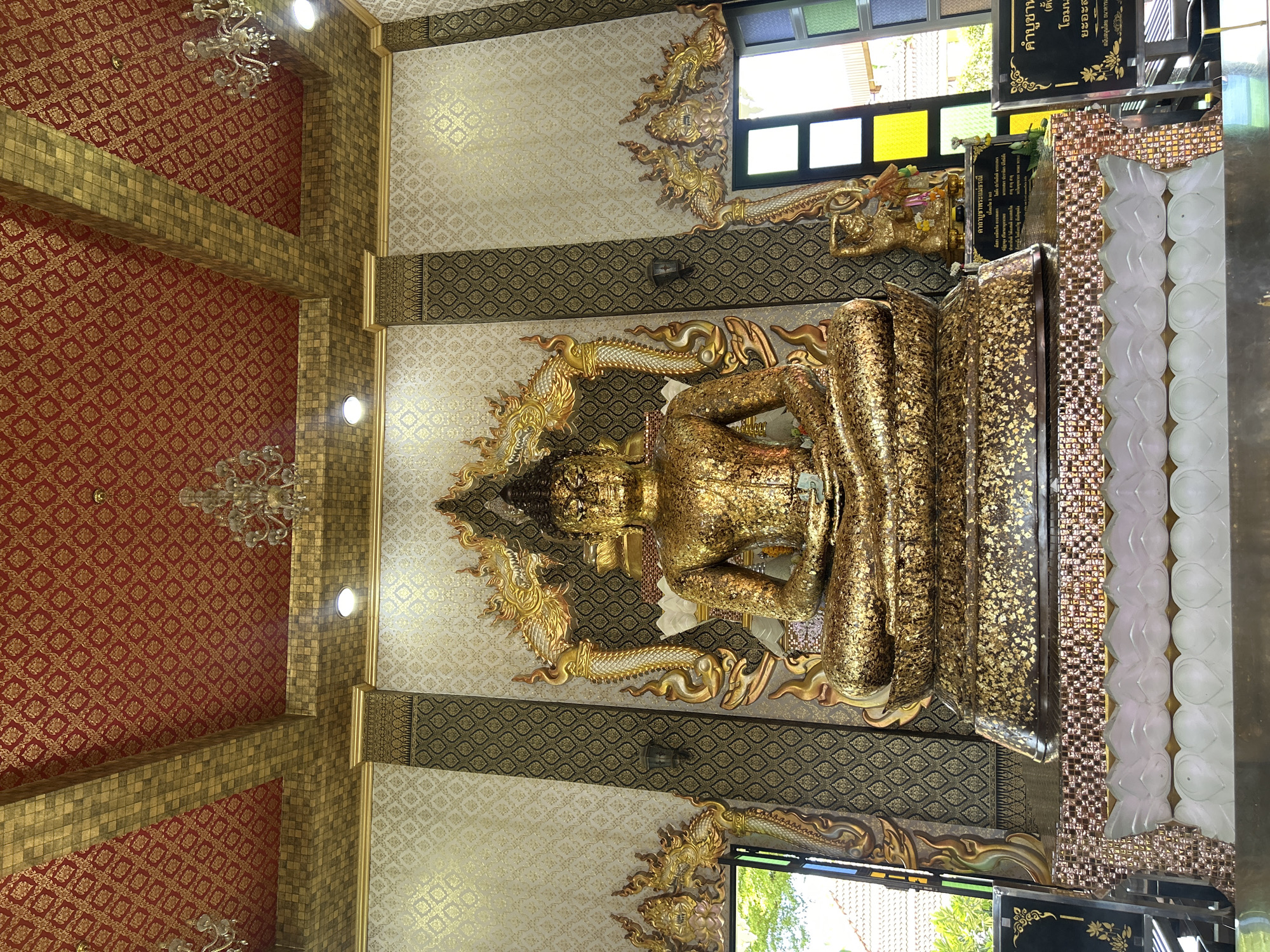 A golden Buddha statue sits centrally in a temple hall, adorned with gold leaf. The statue is seated in a meditative pose on an ornate, tiered base. The hall features elaborate decorations, including a red and gold patterned ceiling with chandeliers, and walls with textured golden and white patterns. Flanking the Buddha statue are decorative golden dragon-like motifs. To the left and right of the hall, open windows reveal glimpses of outdoor greenery and architecture. Small golden figures and religious plaques are visible on the altar in front of the Buddha.