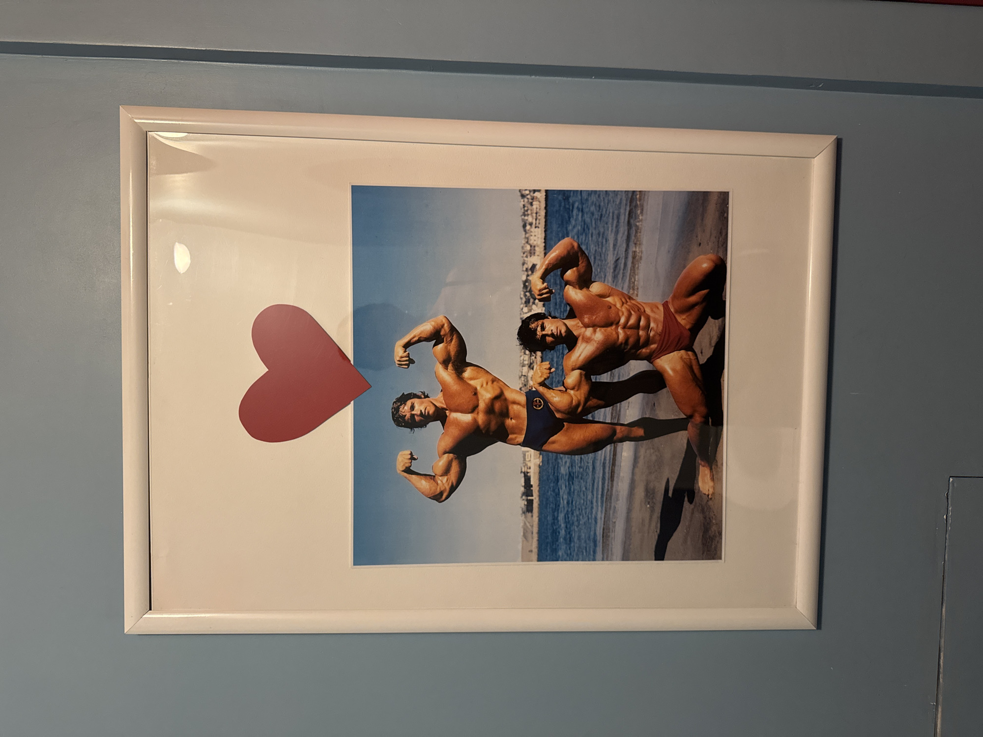 A white framed picture is mounted vertically on a light blue-gray wall. The wall appears smooth with a matte finish. The frame itself is white, rectangular, and has a slightly rounded profile. Visible within the frame, above the primary image, is a piece of white matting. Affixed to the upper central portion of this matting is a solid red heart-shaped cutout.
Below the heart and matting, the main subject is a color photograph depicting two male bodybuilders posing on a beach. The taller bodybuilder, identified as Arnold Schwarzenegger, stands on the left, flexing his biceps and wearing dark blue posing trunks. The shorter bodybuilder, identified as Franco Columbu, kneels on the right, also flexing his biceps and wearing red posing trunks. Both men exhibit highly developed musculature and a tan complexion.
The setting of the photograph is a sandy beach bordering a body of water with small waves, likely an ocean or large sea. In the background, across the water, a cluster of white or light-colored buildings can be seen ascending a hillside under a clear blue sky. Shadows from the men and an unseen object are cast on the sand, suggesting bright daylight. Several reflections from an internal light source are visible on the protective glass of the frame, particularly in the upper right quadrant. The location of the scene depicted in the photograph is Unknown, Unknown.