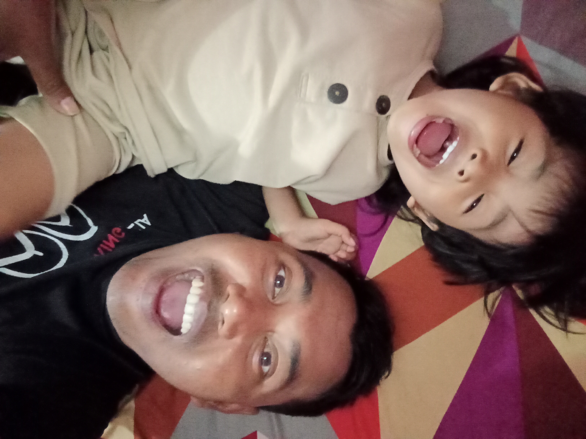 The image shows a close-up, overhead shot of two people lying down, with the image rotated so their heads are towards the bottom. On the right is a young child with dark hair, their mouth wide open in a laugh or shout, wearing a light-colored shirt with two dark buttons. To the left and slightly below the child is an adult, also with dark hair and their mouth open in a smile, revealing teeth. They are wearing a black t-shirt with white lettering that is partially visible. The background appears to be a patterned surface, possibly a blanket or bedspread, with geometric shapes in shades of orange, yellow, pink, and brown. The lighting is direct and bright, creating some glare. This appears to be a casual, personal photograph taken indoors. While the image itself does not provide specific geographic markers for Bloro, Indonesia, it depicts a common domestic scene likely found anywhere.