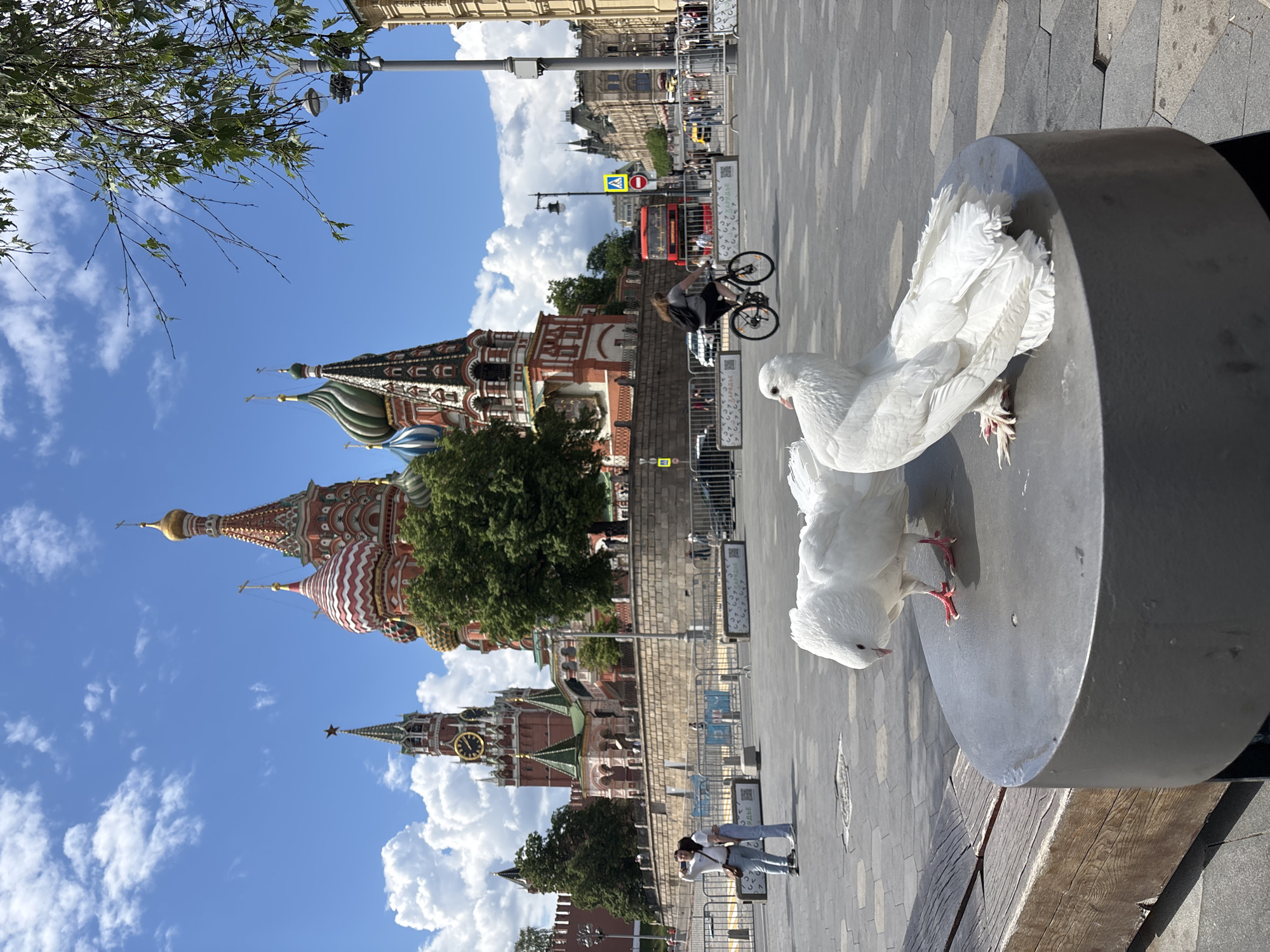 Two white doves perch on a cylindrical concrete structure. In the background is the colorful architecture of St. Basil's Cathedral and the Kremlin Clock Tower, indicating the location is Red Square in Moscow, Russia. People can be seen walking in the background, with one person riding a bicycle. A double-decker tour bus is visible further back. The sky is blue with fluffy white clouds. Trees are seen among the buildings.