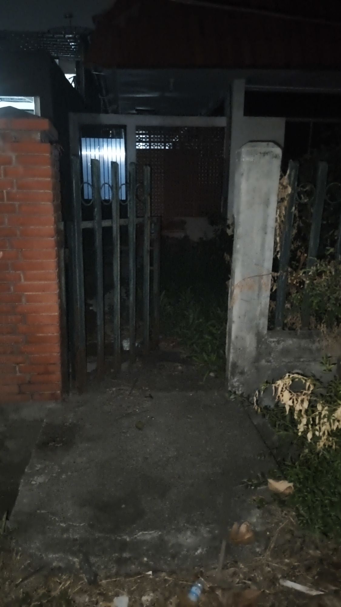 This image depicts a dimly lit pathway leading to a gated entrance, likely in South Jakarta, Indonesia. The pathway is paved with rough concrete and is bordered by overgrown vegetation on the right. To the left, a brick wall is visible, and to the right, a large concrete pillar supports a gated structure. The gate itself is made of dark metal bars. Beyond the gate, a building with a white patterned screen is visible, illuminated by interior lights. Debris, including dry leaves and a plastic bottle, is scattered on the ground. The overall scene suggests an outdoor area, possibly a residential or commercial property, at night or in low light conditions.