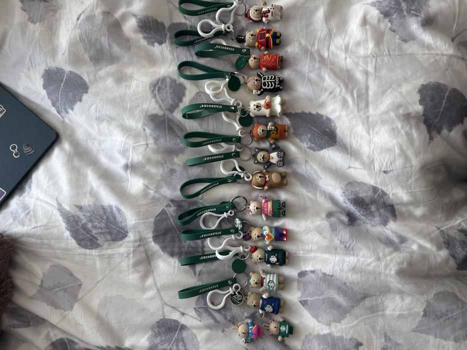 A collection of Starbucks bear keychains arranged in a vertical line on a patterned bedspread. The bears feature various costumes and themes, each attached to a green Starbucks-branded loop and a white clasp. A laptop with stickers is visible in the upper left corner. The bedspread has a gray and white floral leaf design.