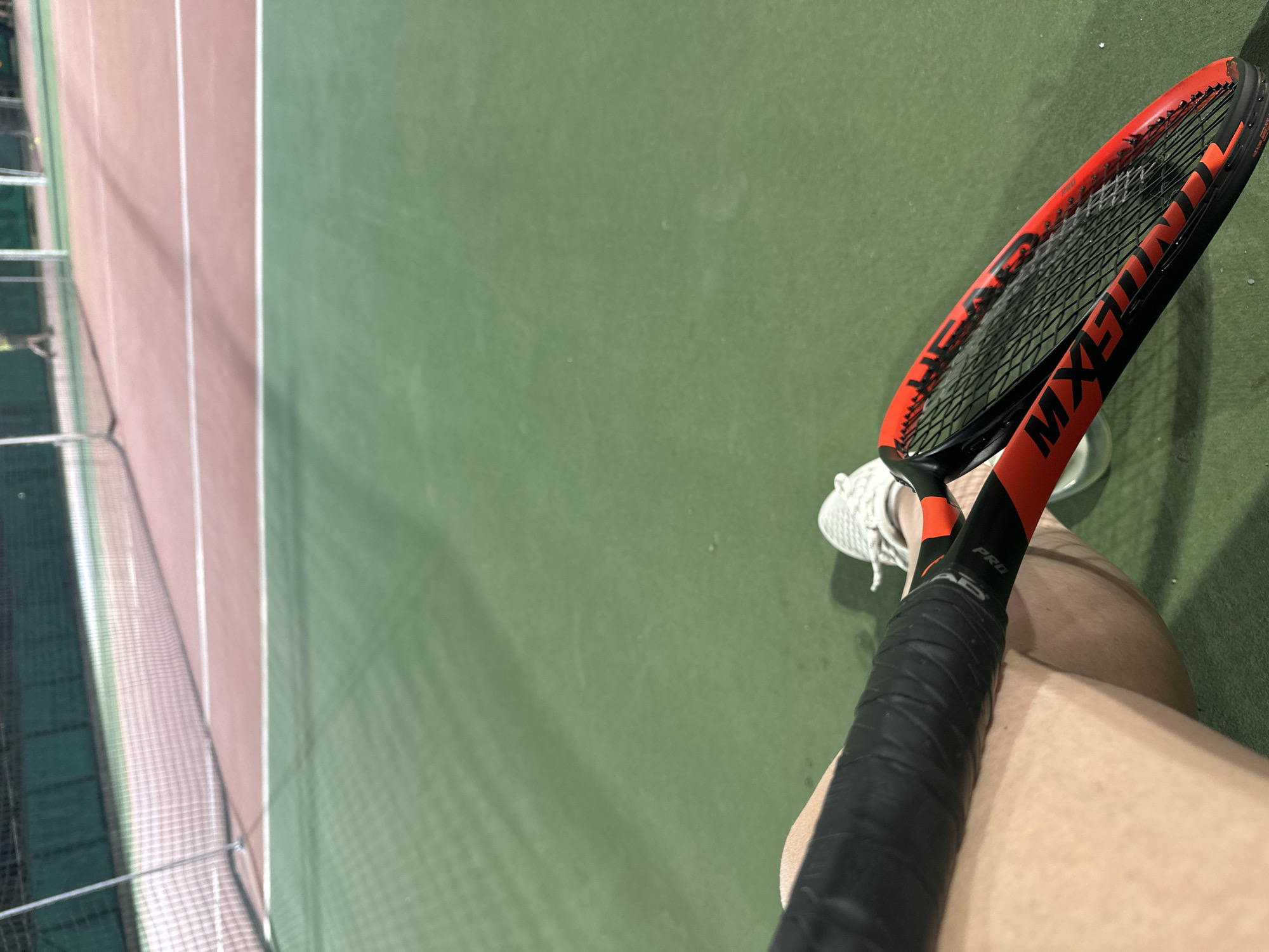 This is a first-person perspective image taken at a tennis court. The foreground is dominated by a tennis racket with a black grip and a bright orange frame, held by an unseen person whose legs are visible. The person is wearing a white tennis shoe. In the background, a tennis net divides the court, which has a green surface. Beyond the net, a portion of a pink-colored court is visible. The lighting suggests it is either evening or an indoor court. The location is identified as a tennis court in the city of Sofia, Bulgaria.