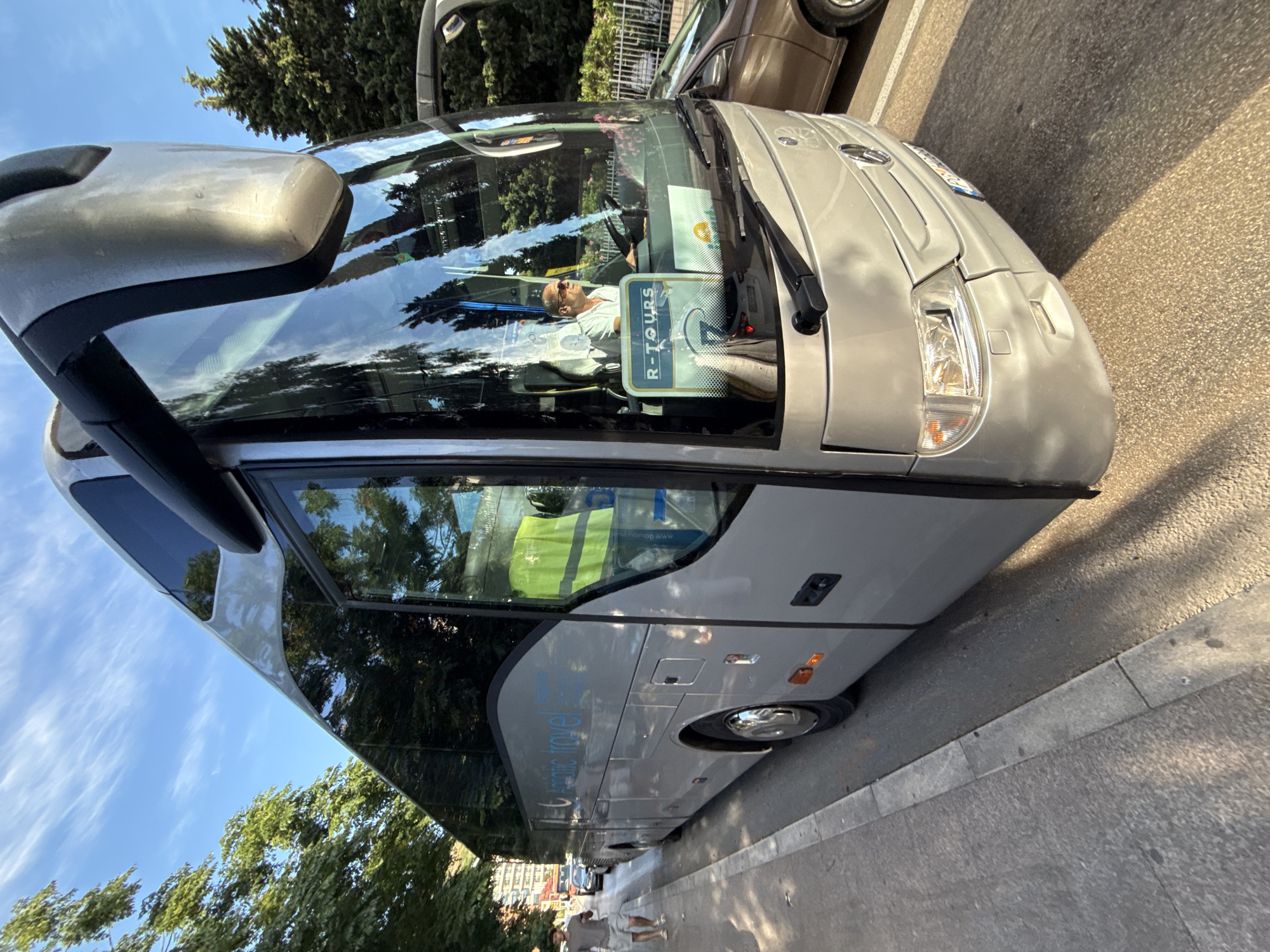 A silver bus with the logo "Adriatic Travel" is parked on the side of the road in Budva, Montenegro. The bus is facing towards the right of the frame and its front is partially visible. A man in a white shirt and sunglasses is driving the bus, and a sign with "R-TOURS" is displayed on the dashboard. The bus is on a street with trees and buildings in the background. A brown car is parked next to the bus on the right side of the frame. A person in a grey t-shirt and shorts is walking on the sidewalk on the left side of the frame. The sky is blue with scattered clouds. The scene appears to be in the afternoon, with sunlight casting shadows on the ground.