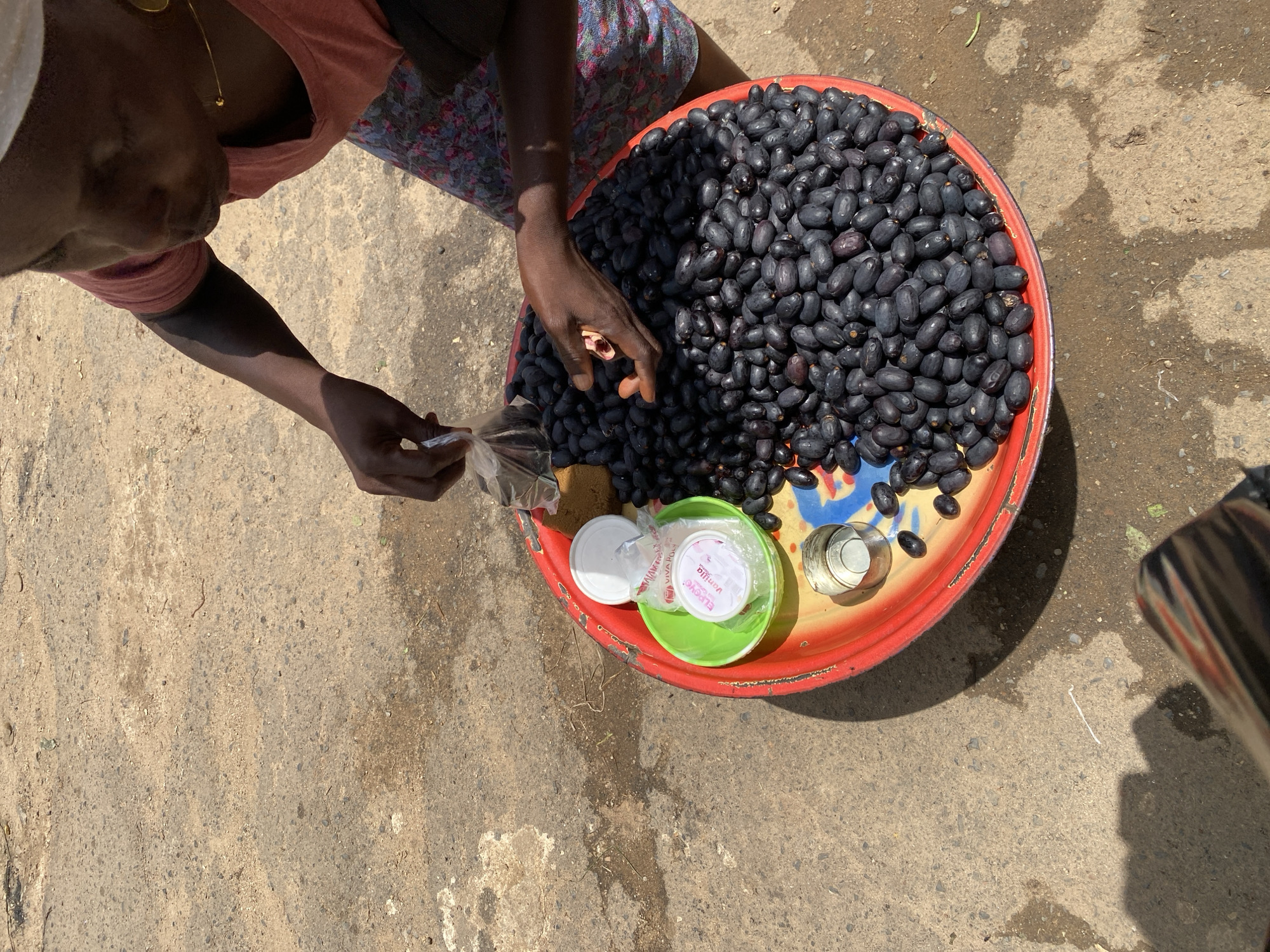 In Jos, Nigeria, a person is seen interacting with a large red tray filled with dark purple, elongated fruits, possibly Java plums. The tray also contains a small green bowl with two white containers, one of which has a pink label with "VIVA FOOD Vanilla" printed on it. The person's hands are visible, with one hand reaching into a clear plastic bag and the other delicately holding one of the fruits. The ground beneath the tray appears to be a dusty, uneven surface, consistent with an outdoor market setting in a city. The lighting suggests it is daytime, with shadows cast by the person and the tray.