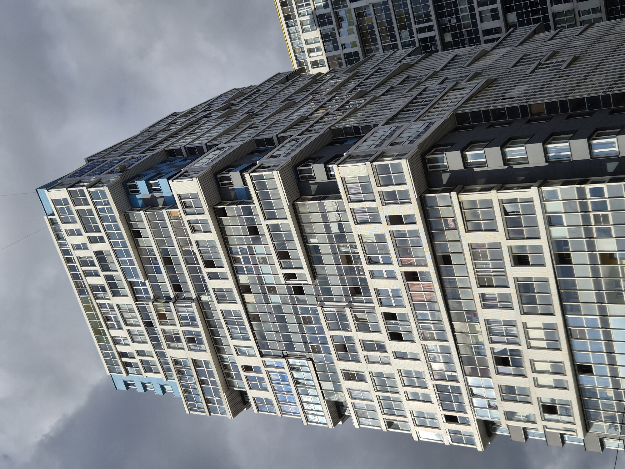 A low-angle, full-shot captures a cluster of modern apartment buildings against a dramatic, overcast sky. The primary building, a tall, multi-faceted structure with a staggered facade, dominates the foreground. Its exterior is a mosaic of light-colored panels and numerous large windows, many of which reflect the muted sky. Distinctive blue accents are visible on upper levels and balconies. To the right, another similarly constructed, though less prominent, building shares the frame, its facade also composed of windows and lighter exterior panels. The overall impression is one of contemporary urban architecture under a moody, cloudy atmosphere.