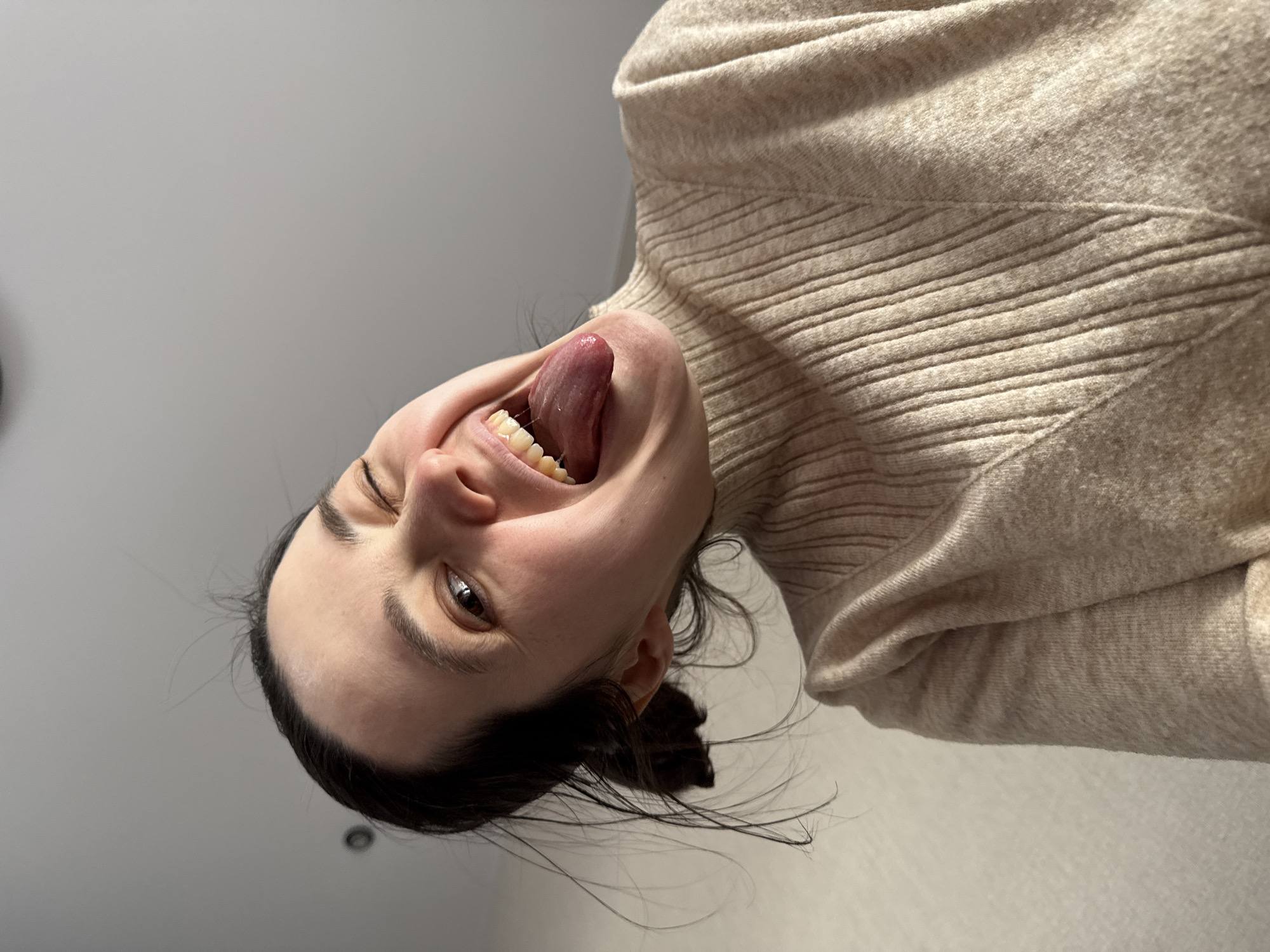 A woman is looking down at the camera, sticking her tongue out. She is wearing a beige sweater with a ribbed texture. Her hair is dark and pulled back. The background is a plain white ceiling. There is no indication of the location being Izhevsk, Russia.