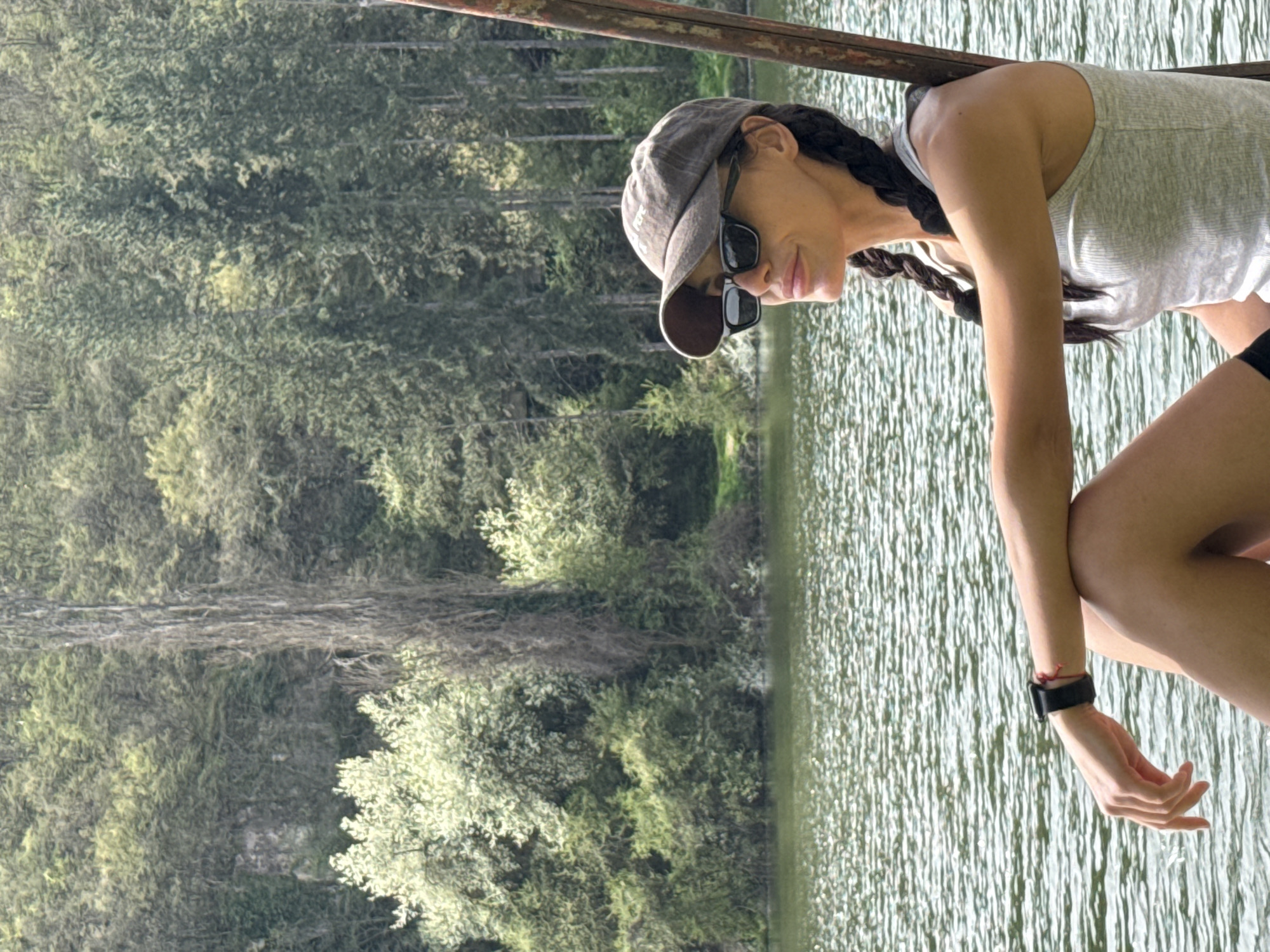 A woman wearing sunglasses and a cap leans against a metal railing by a body of water reflecting trees. The environment appears to be natural, suggesting a park or lake area. It is not possible to confirm the location as Pleven, Bulgaria based on the image alone.