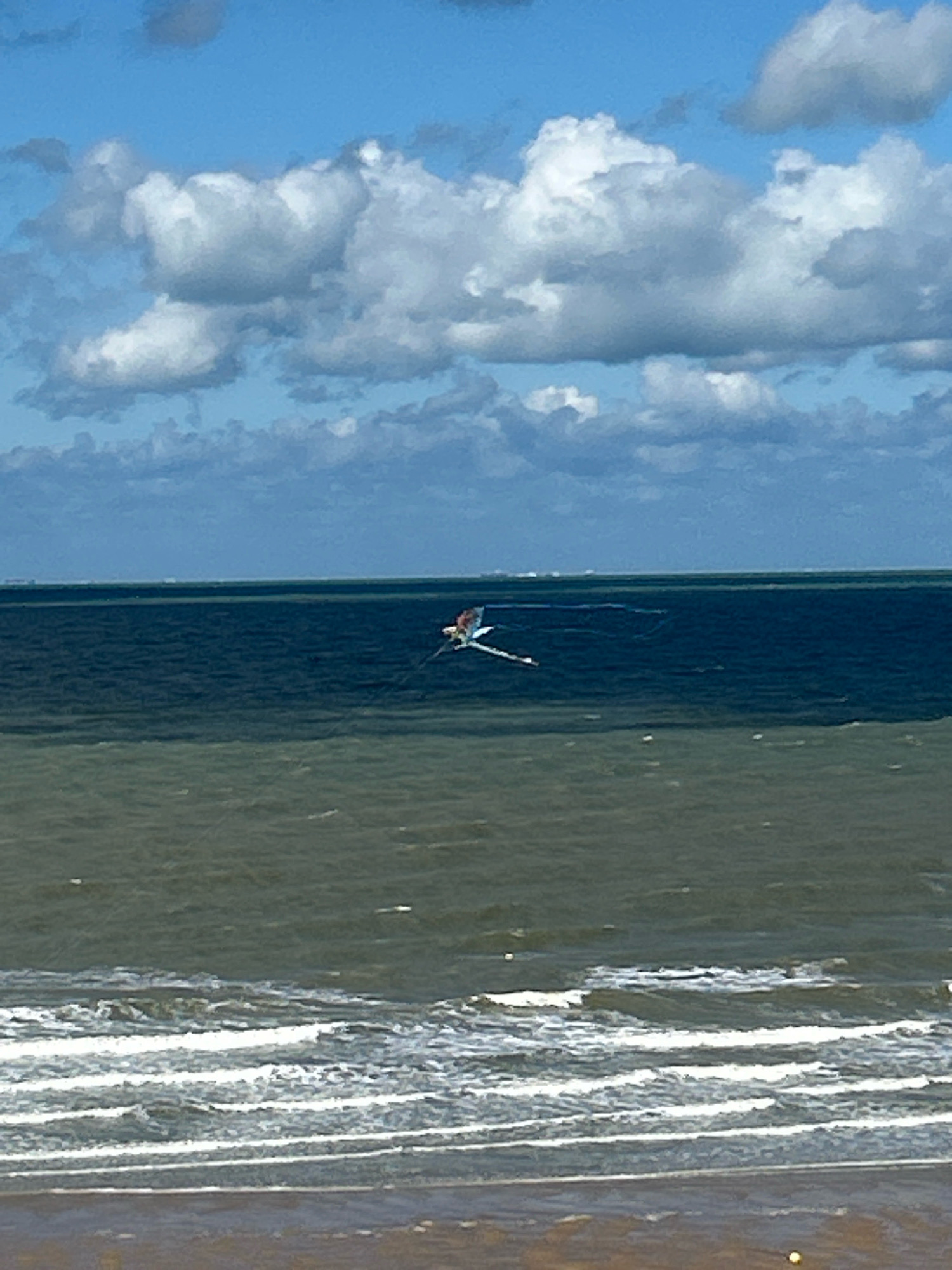 A flamingo kite is flying over the North Sea off the coast of Ostend, Belgium. The kite, which is predominantly white with pink and red accents, is in mid-flight, with its wings outstretched. The kite is positioned in the upper middle section of the image, against a backdrop of a bright blue sky filled with fluffy white clouds. The sea is dark blue and calm, with a distinct line separating it from the lighter, greenish-brown water closer to the shore. The foreground shows the sandy beach and some small waves breaking on the shore.