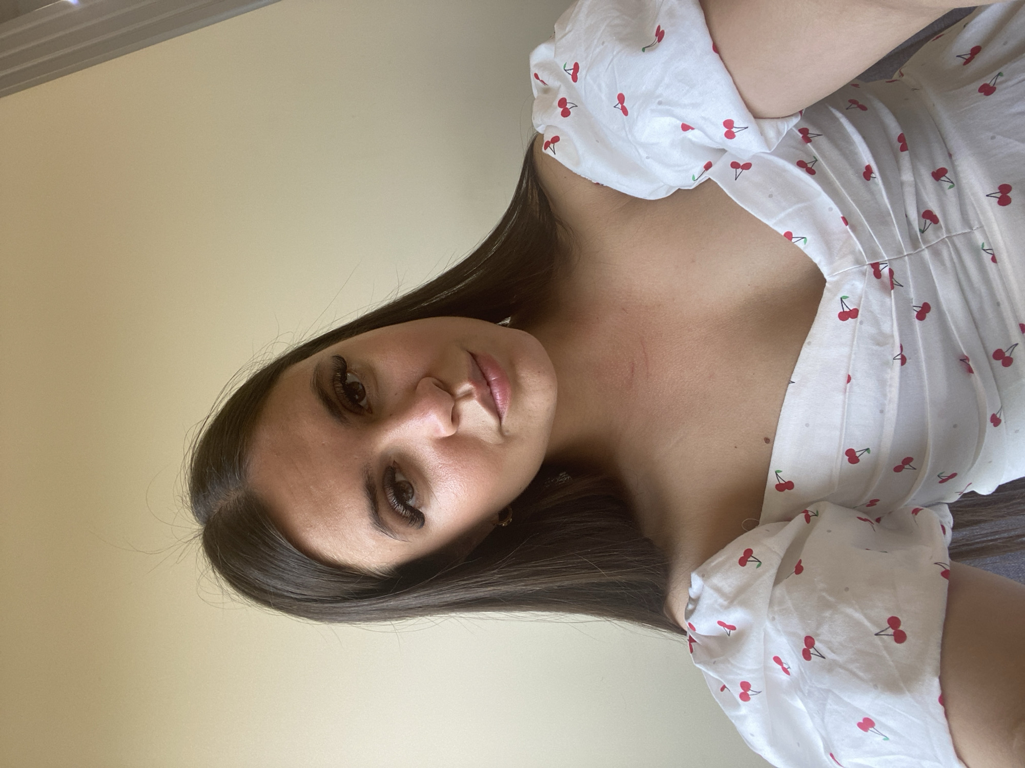 A young woman with long, dark hair is looking at the camera. She is wearing a white dress with a cherry print. The background is a plain wall. There is no indication of location within the city of Derbent, Russia.