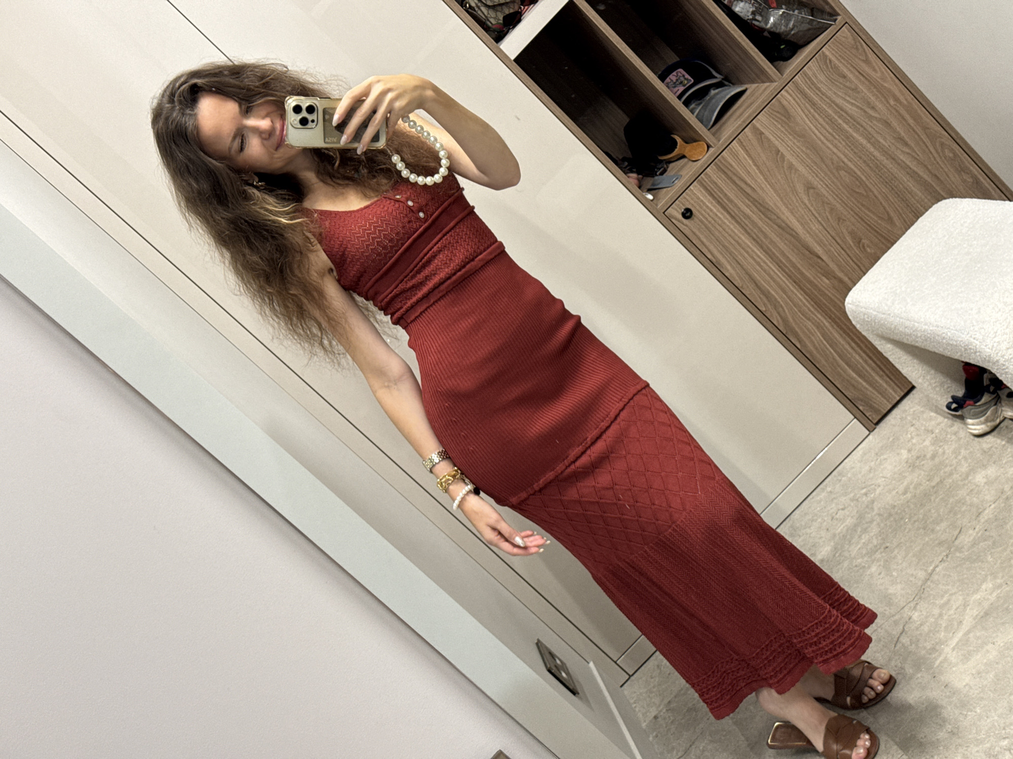 A woman is taking a mirror selfie in what appears to be a modern, minimalist closet or dressing area. She is wearing a long, red, knitted dress with a textured pattern and a low neckline, accessorized with a pearl necklace and several bracelets on her left wrist. She is also wearing brown open-toed sandals. Her hair is long and wavy, and she is smiling at her reflection. In the background, there is a tall white wardrobe with wooden accents, featuring shelves filled with various items, including bags, hats, and other accessories. The flooring is a light-colored, textured material, possibly marble or stone. The lighting is bright, suggesting an indoor setting.