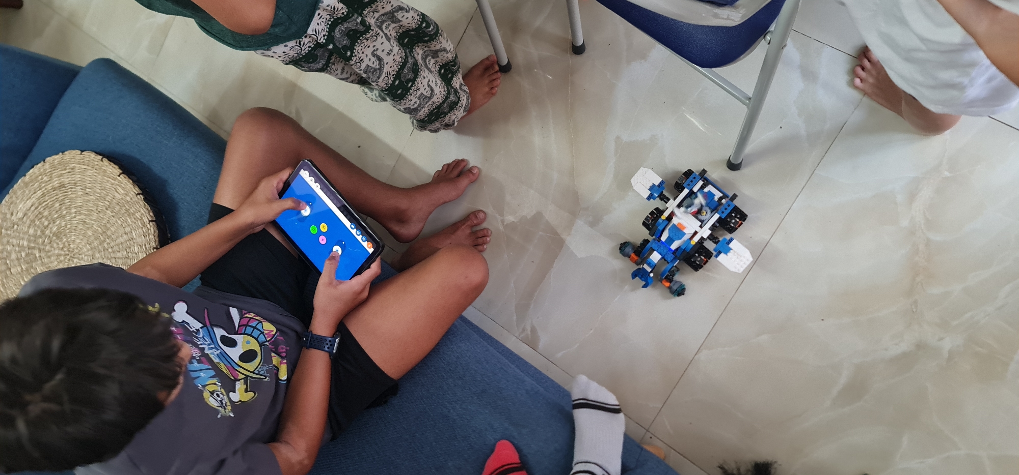 An overhead shot depicts an indoor scene with multiple individuals. A child sits on a blue couch, engrossed in a tablet with a game displayed. The child wears a grey t-shirt with a skull and crossbones design and black shorts. To the child's left sits a round, woven placemat on the couch.
Adjacent to the couch, a white, tiled floor is visible. Near the child playing the game is a blue and white LEGO vehicle. Parts of other individuals are visible, including feet, legs wearing patterned green and white pants, a leg in white shorts, and feet wearing socks with red and white stripes. A portion of a blue chair frame is also visible.
There is no information to indicate where this picture was taken, including the city of Đà Nẵng, Vietnam.