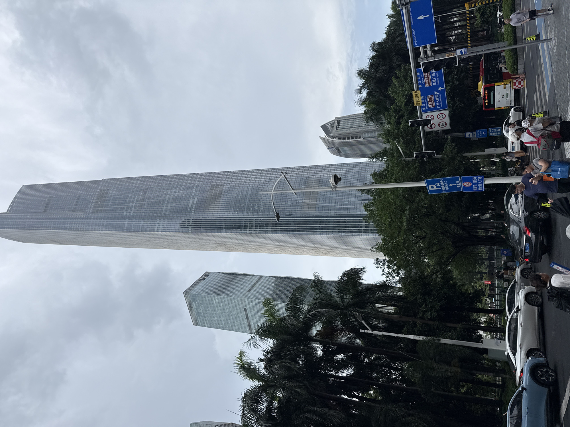 The image depicts a street scene in Tianhe District, Guangzhou, China, dominated by a very tall, modern skyscraper with a glass facade. Several other modern buildings are visible in the background, some with glass exteriors and one with a distinctive curved, pointed top. Palm trees and other lush green foliage are present in the foreground and between buildings, suggesting a tropical or subtropical urban environment.
The street is busy with traffic, including several cars, a bus, and a taxi. Pedestrians are crossing a street at a crosswalk. Traffic signals and directional signs are prominently displayed, including signs in Chinese characters and English, indicating destinations like "Zhujiang East Rd", "Linjiang Ave", and "Guangzhou 2nd Children's Hospital". The sky is overcast, suggesting a cloudy day. One person is seen holding an umbrella, which may indicate light rain or protection from the sun. The overall impression is of a bustling, modern commercial district within a large Chinese city.