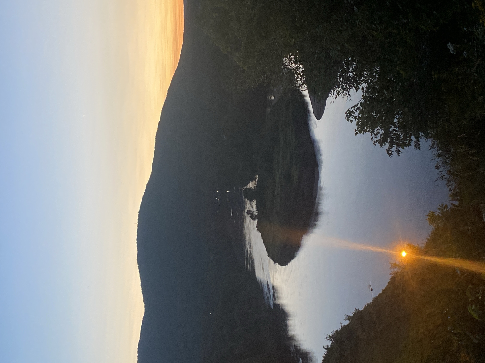 This is a photograph taken at dusk overlooking a wide, winding river bordered by forested hills. The sky transitions from a clear pale blue at the top to a warm orange hue near the horizon, indicating the setting sun. The sun itself is visible low in the frame on the left, its bright light reflecting strongly on the water and creating a lens flare that extends upwards. The river curves through a valley, with a prominent island in the center of the bend. The surrounding landscape is covered in dense, dark green foliage, suggesting a natural, undeveloped area. In the distance, faint lights can be seen on the far bank of the river, implying the presence of a settlement or buildings. The overall impression is one of natural beauty and tranquility at twilight. The location is likely within or near Saint-Gabriel-de-Valcartier, Canada, a region known for its scenic river valleys and surrounding natural landscapes.