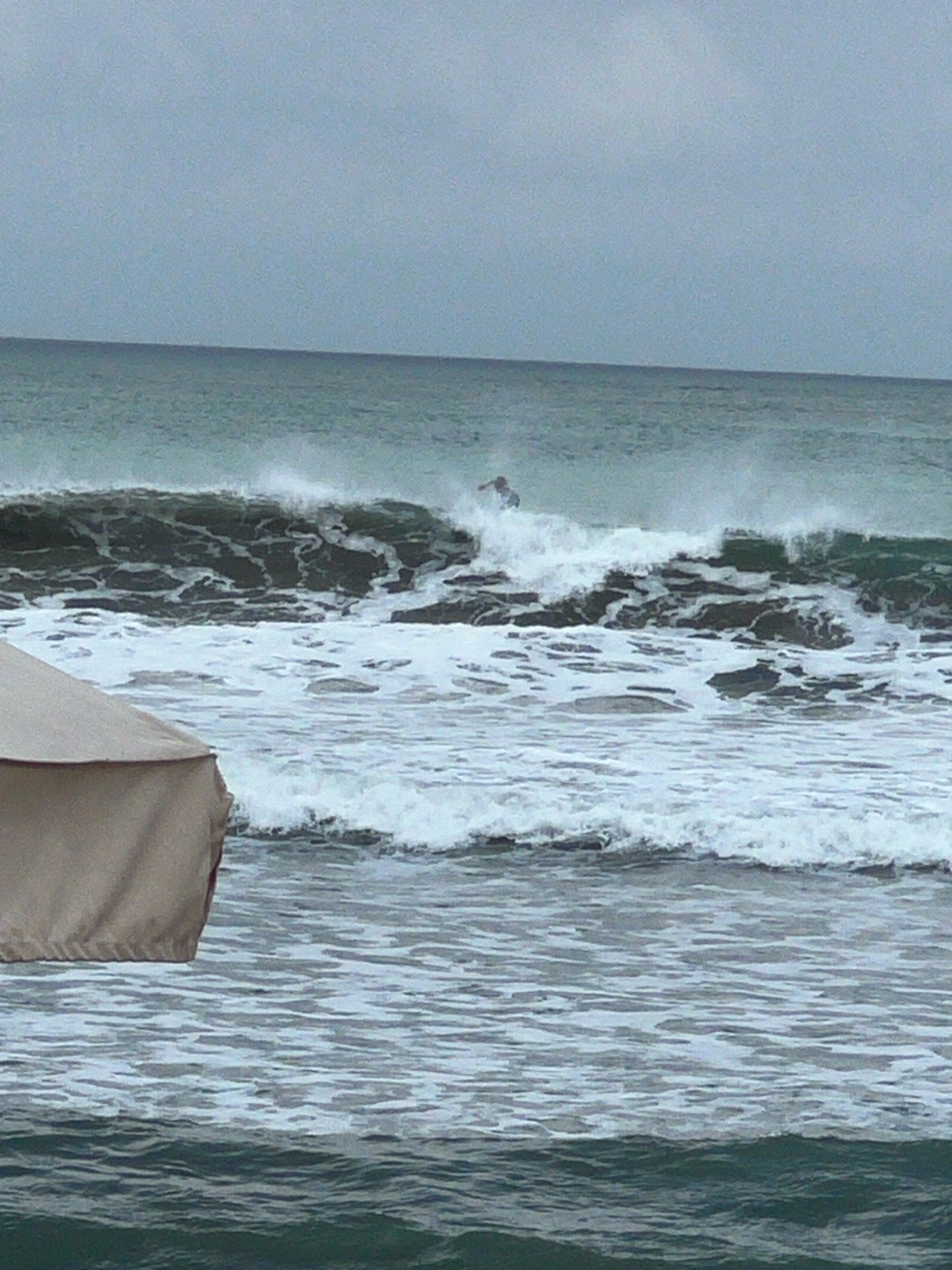 The image depicts a surfer riding a wave in the ocean off the coast of Kuta, Indonesia. The sky is overcast, and the water is choppy with white foam. In the foreground, a portion of a beach umbrella is visible, suggesting the presence of a beachside setting. The surfer is in the middle distance, actively engaged in surfing the breaking wave. The environment is typical of a coastal area with the ocean and sky as prominent features.
