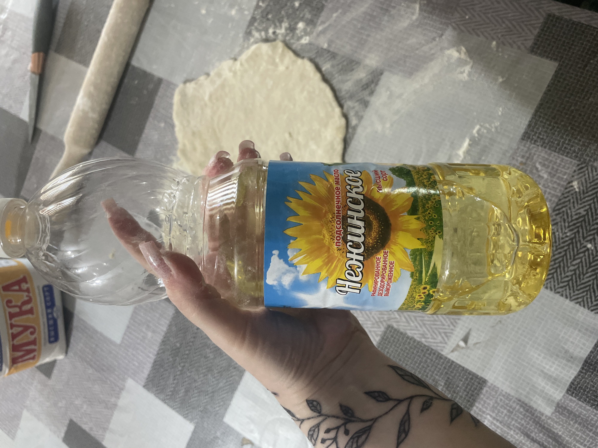 The image shows a person's hand holding a bottle of "Нежинское" (Nezhinskoe) sunflower oil. The hand has long, light-colored artificial nails and a tattoo of leaves on the wrist and forearm. The bottle of sunflower oil is clear plastic with a blue label featuring a large sunflower and fields of sunflowers. Text on the label reads "подсолнечное масло" (sunflower oil), "Нежинское" (Nezhinskoe), and "Высший сорт" (Highest grade), as well as "РАФИНИРОВАННОЕ ДЕЗОДОРИРОВАННОЕ ВЫМОРОЖЕННОЕ" (refined, deodorized, and winterized).
The setting appears to be a kitchen or dining area, with a grey and white checkered tablecloth visible. In the background, there is a rolling pin, a partially rolled-out piece of dough, and a knife. A package labeled "МУКА" (flour) is also partially visible on the left side of the image. The scene suggests the preparation of food, likely baking or cooking. There are no specific cues about the time of day or weather. The overall mood is neutral, focusing on the task at hand.