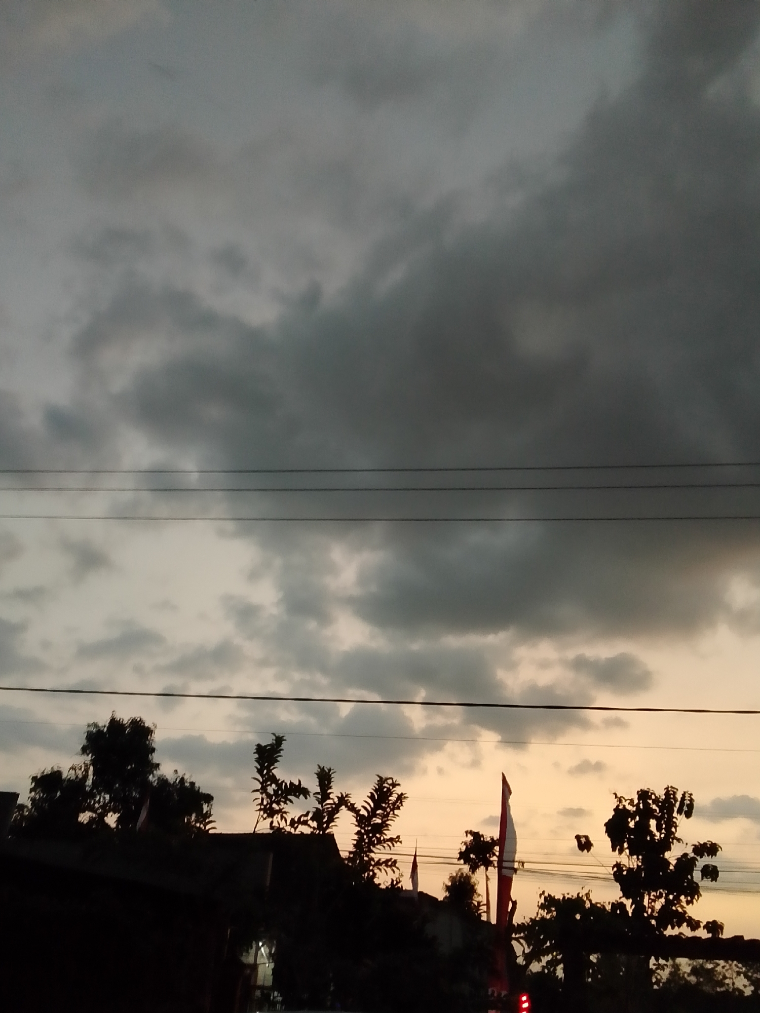 A low-angle shot captures a cloudy sky with a gradient of grays and pale oranges, suggesting either dawn or dusk. Three horizontal power lines are visible across the frame. Silhouetted against the sky in the lower portion of the image are the tops of trees and a building. Among the trees, two Indonesian flags are visible, one a prominent red and white banner. The scene appears to be outdoors, possibly in a residential or semi-rural area given the presence of trees and a building. The overall atmosphere is subdued due to the low light and overcast conditions. The location is identified as Mojotengah, Indonesia.