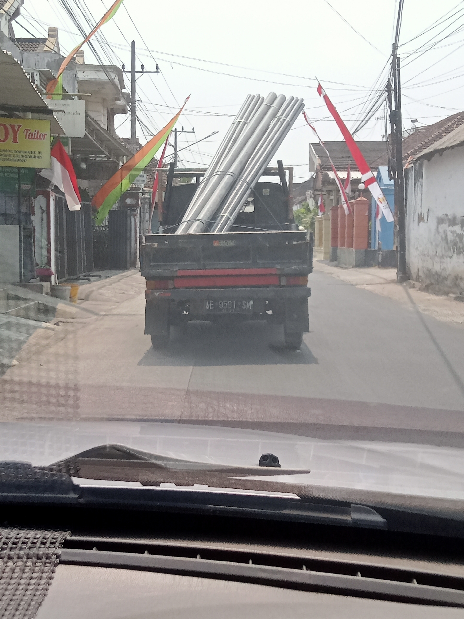 In Mangunsari, Indonesia, a flatbed truck loaded with large metal pipes travels down a paved street lined with buildings. The truck's license plate reads "AE 9591 SM." Red and white Indonesian flags and a green, orange, and red flag are displayed on poles along the street. The scene is viewed from inside a vehicle, looking through the windshield and over the dashboard.