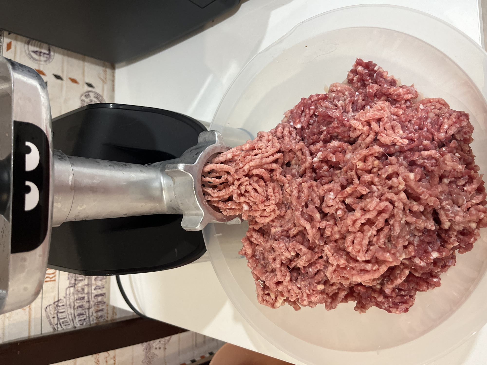 The image shows a meat grinder in operation, processing raw meat into a pile of ground meat in a transparent bowl. The meat grinder is silver and black with a humorous face drawn on the top. The background appears to be a kitchen or food preparation area with tiled walls featuring images of Roman architecture and decorative stamps. The scene is well-lit, focusing on the meat grinding process. There is no visible indication of the location being in Chaikovsky, Russia.
