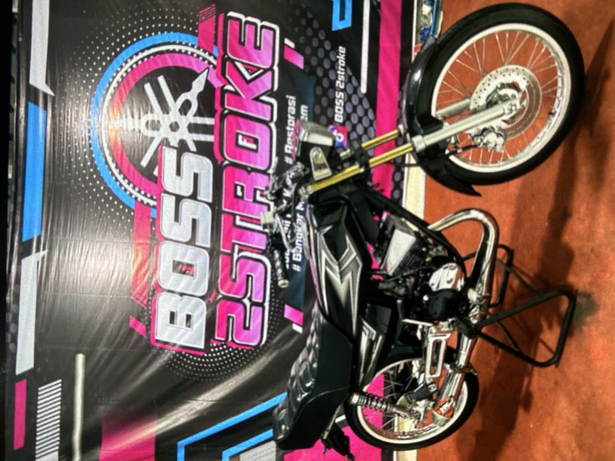 A close-up, vertically oriented shot shows a motorcycle displayed next to a banner in Lhokseumawe, Indonesia. The motorcycle, prominently featured, has a black front fender and silver wire-spoked wheels with chrome rims. The handlebars are gold-colored. Behind the motorcycle is a black banner with pink and white lettering that says "BOSS 2STROKE" in a stylized font. A white symbol is visible in the upper left of the banner. Hashtags and smaller text are also present on the banner. The background is blurry and indistinct, suggesting a close proximity to the objects in focus.